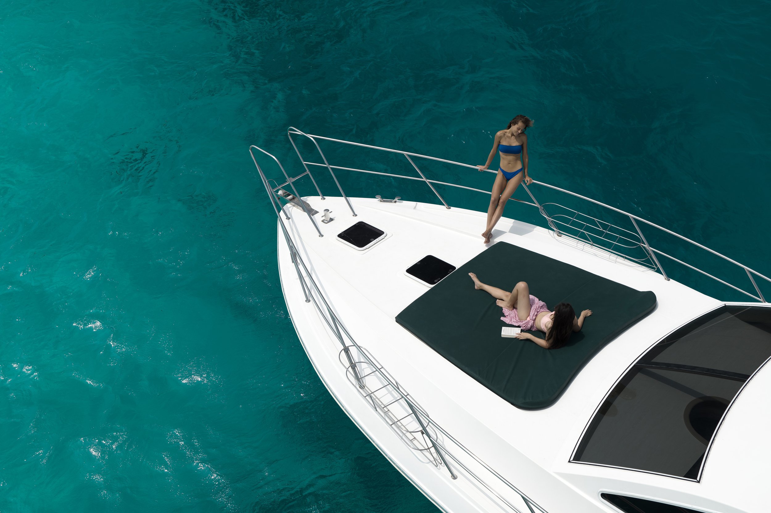 A white yacht floating on turquoise water with two women on board; one lying on a sunbed reading a book, and the other standing near the bow holding a rope, wearing a blue bikini.