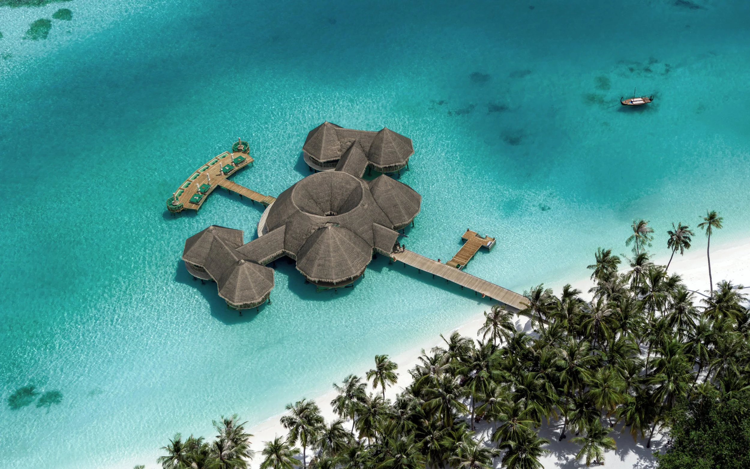 Aerial view of overwater bungalows on a turquoise sea with a sandy beach and palm trees.