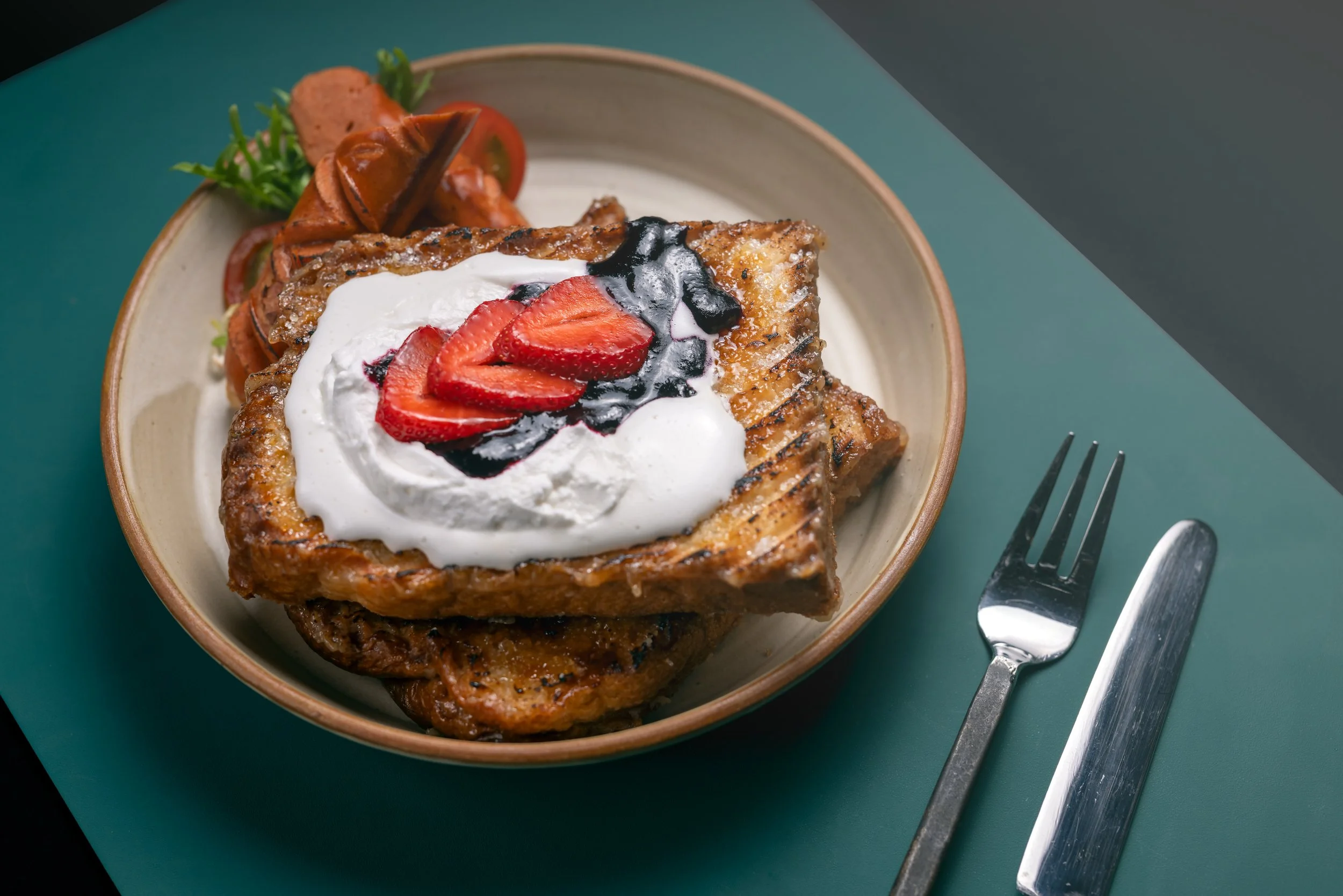 A dessert with a crispy pastry square topped with strawberries, whipped cream, and blueberry sauce, served in a beige bowl on a teal table with a fork and knife beside it.