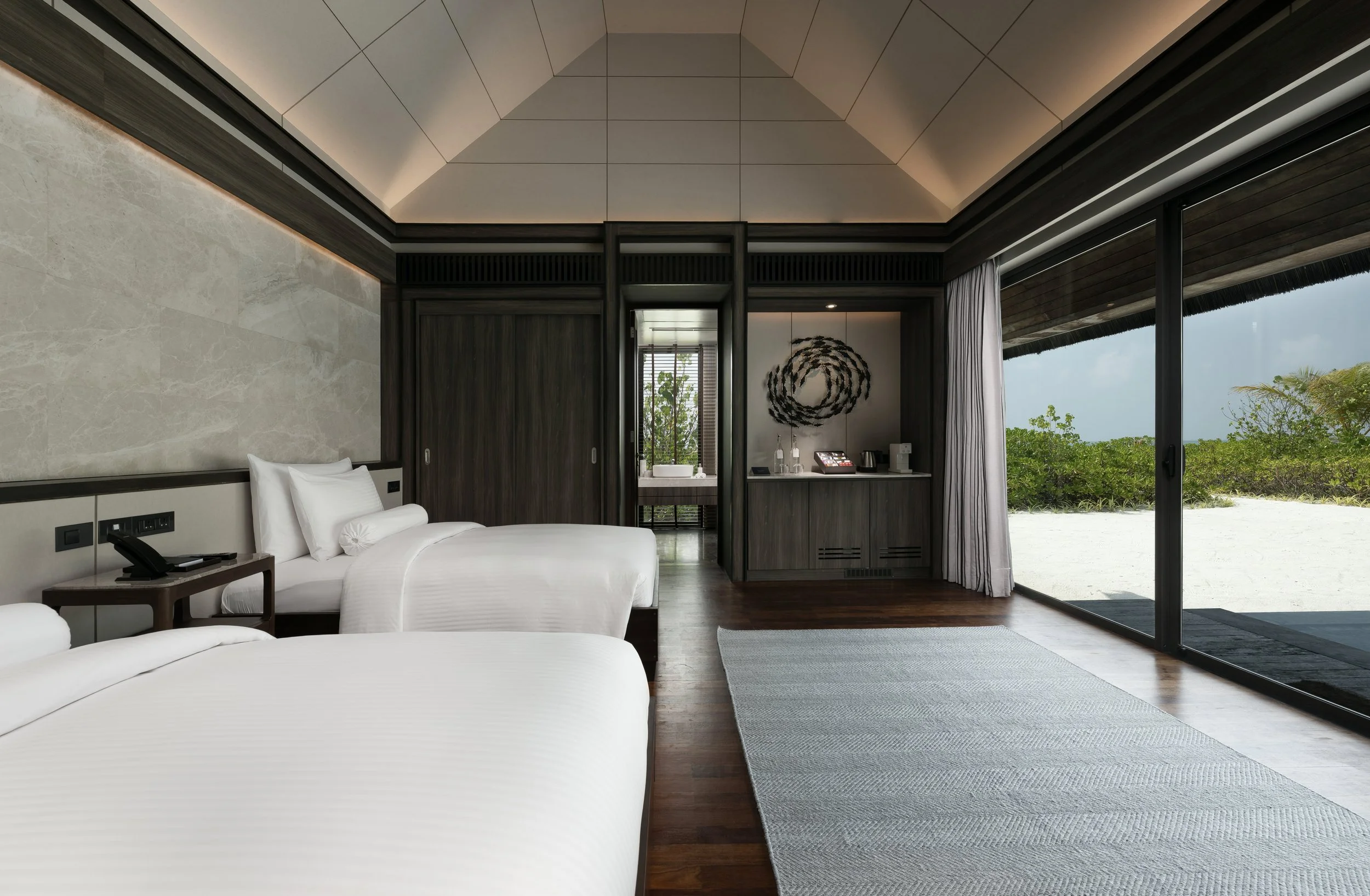 A modern hotel room with white beds, a nightstand with a telephone, and floor-to-ceiling glass doors open to a sandy beach and greenery outside.