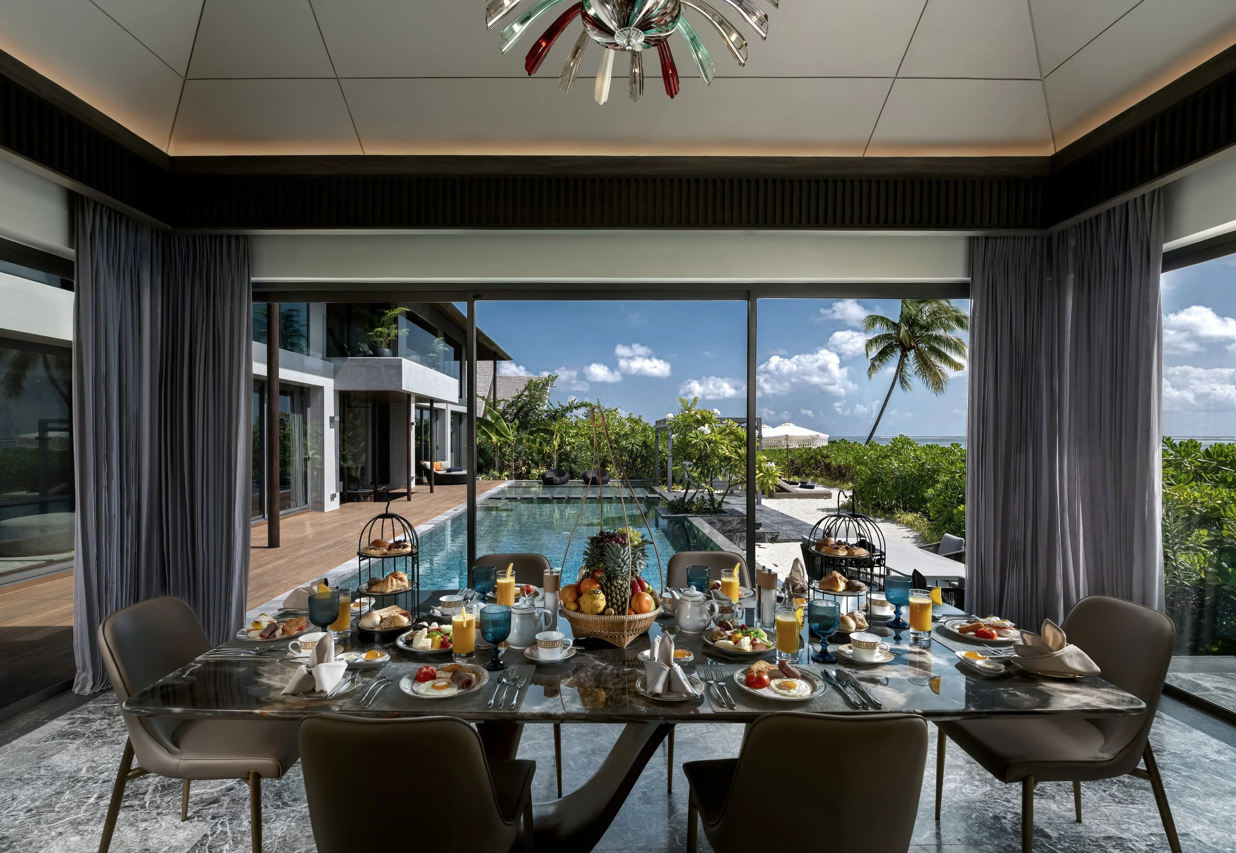 Indoor dining area with a round marble table set for breakfast, including plates with eggs, fruit, croissants, and drinks. Large sliding glass doors open to an outdoor pool and tropical garden with palm trees, overlooking a clear blue sky.
