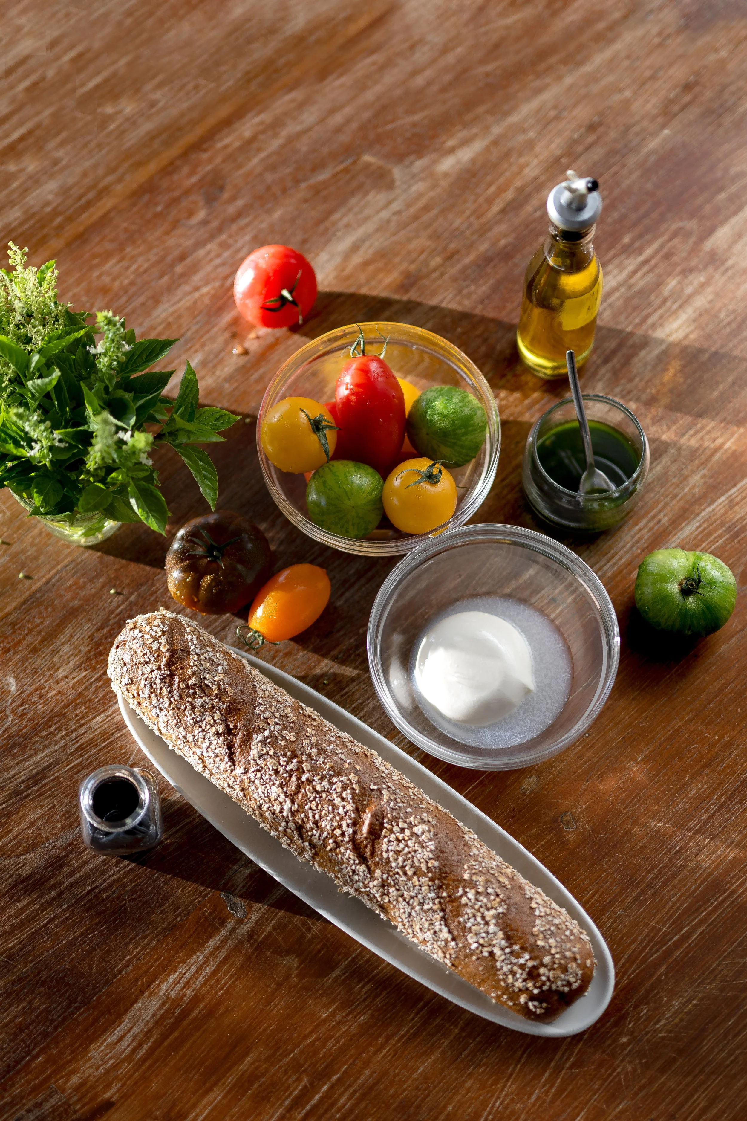 A long, crusty loaf of bread on a white platter, surrounded by small bowls of olive oil, balsamic vinegar, and yogurt, with a bowl of mixed heirloom tomatoes, fresh herbs, and a sprig of basil on a wooden table.