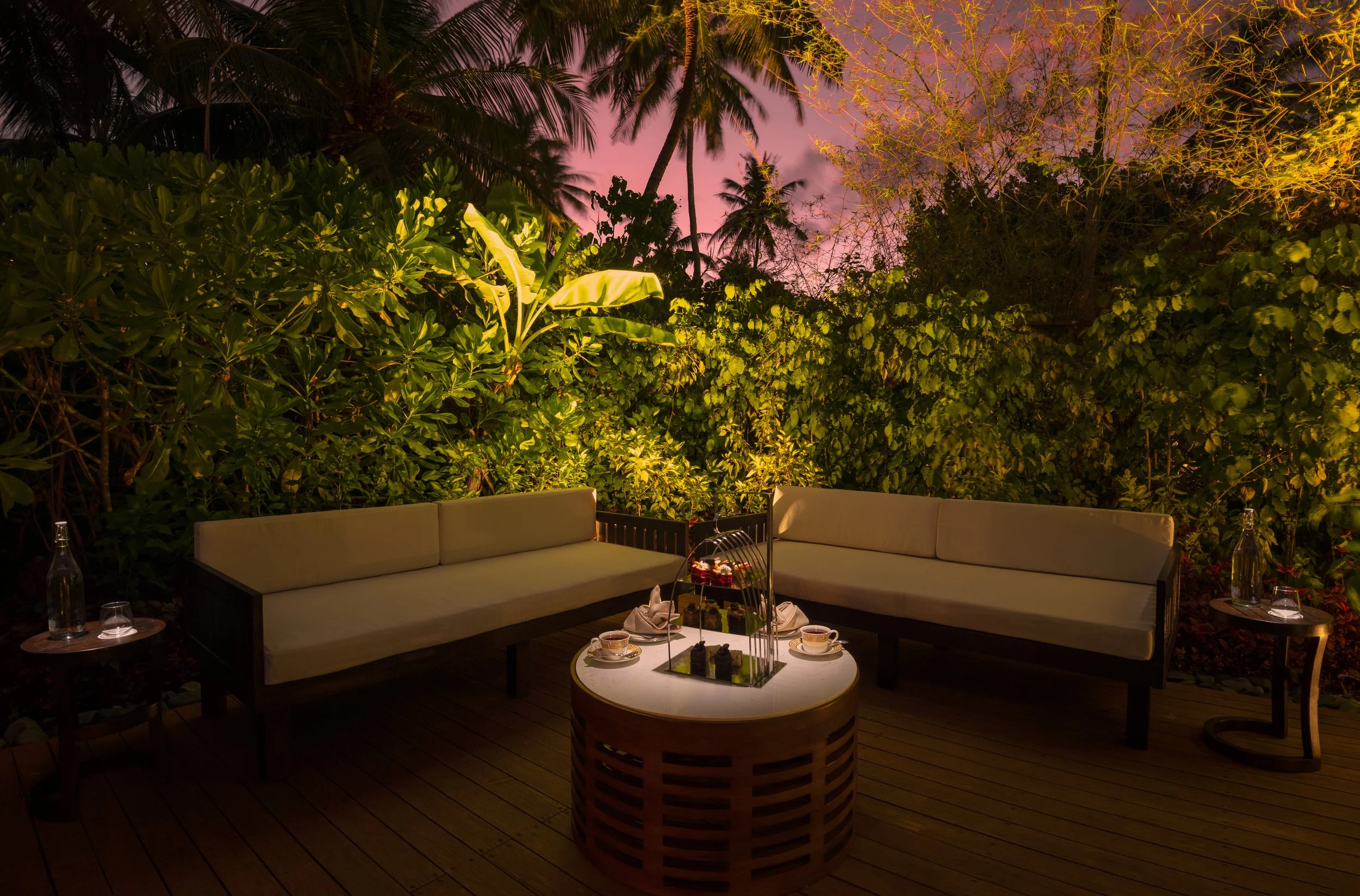 Outdoor patio with beige cushioned seating, surrounded by lush green plants and trees, illuminated by warm lighting at dusk, pink and purple sky visible in the background.