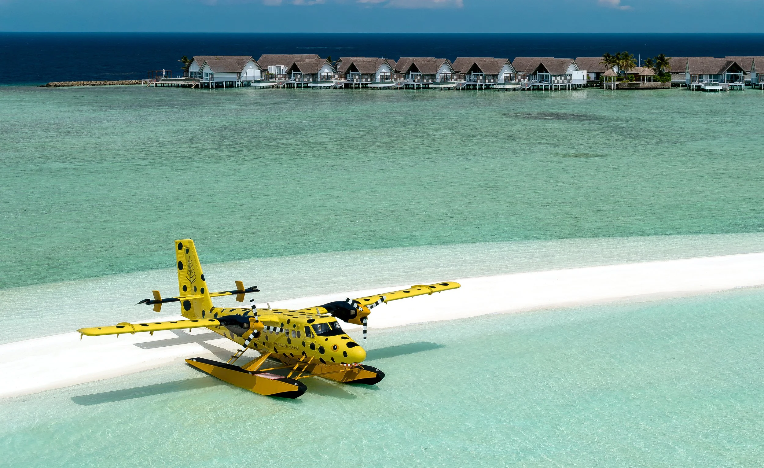 Yellow toy helicopter on a white beach with clear turquoise water and overwater bungalows with thatched roofs in the background, and a dark blue ocean under a partly cloudy sky.