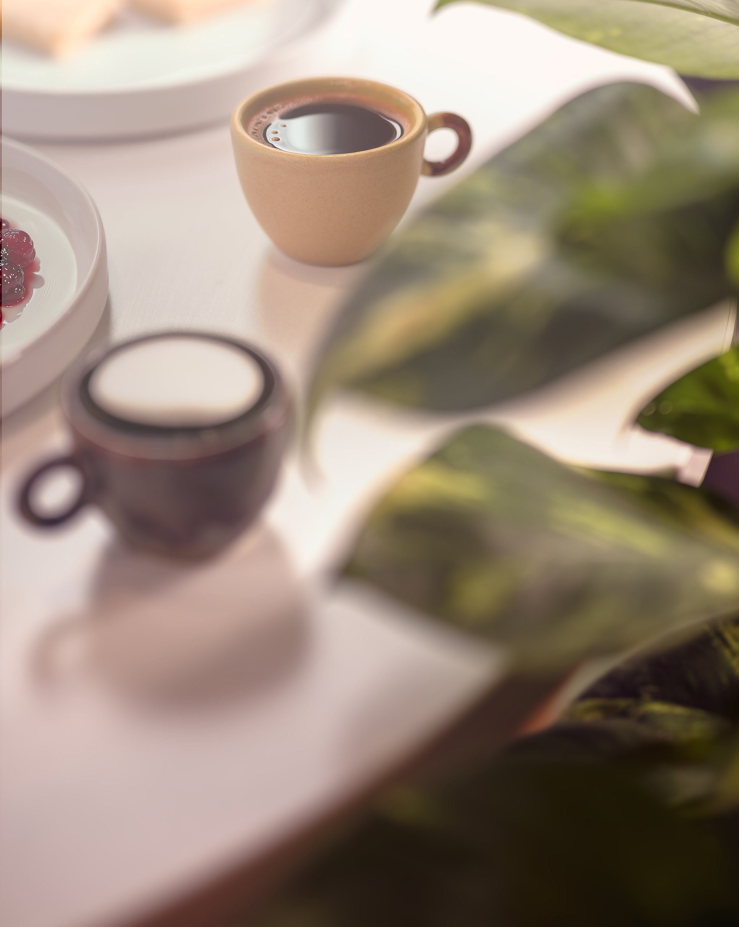 A beige ceramic cup filled with black coffee on a white table, surrounded by plates of food and green leaves, with soft lighting and blurred background.