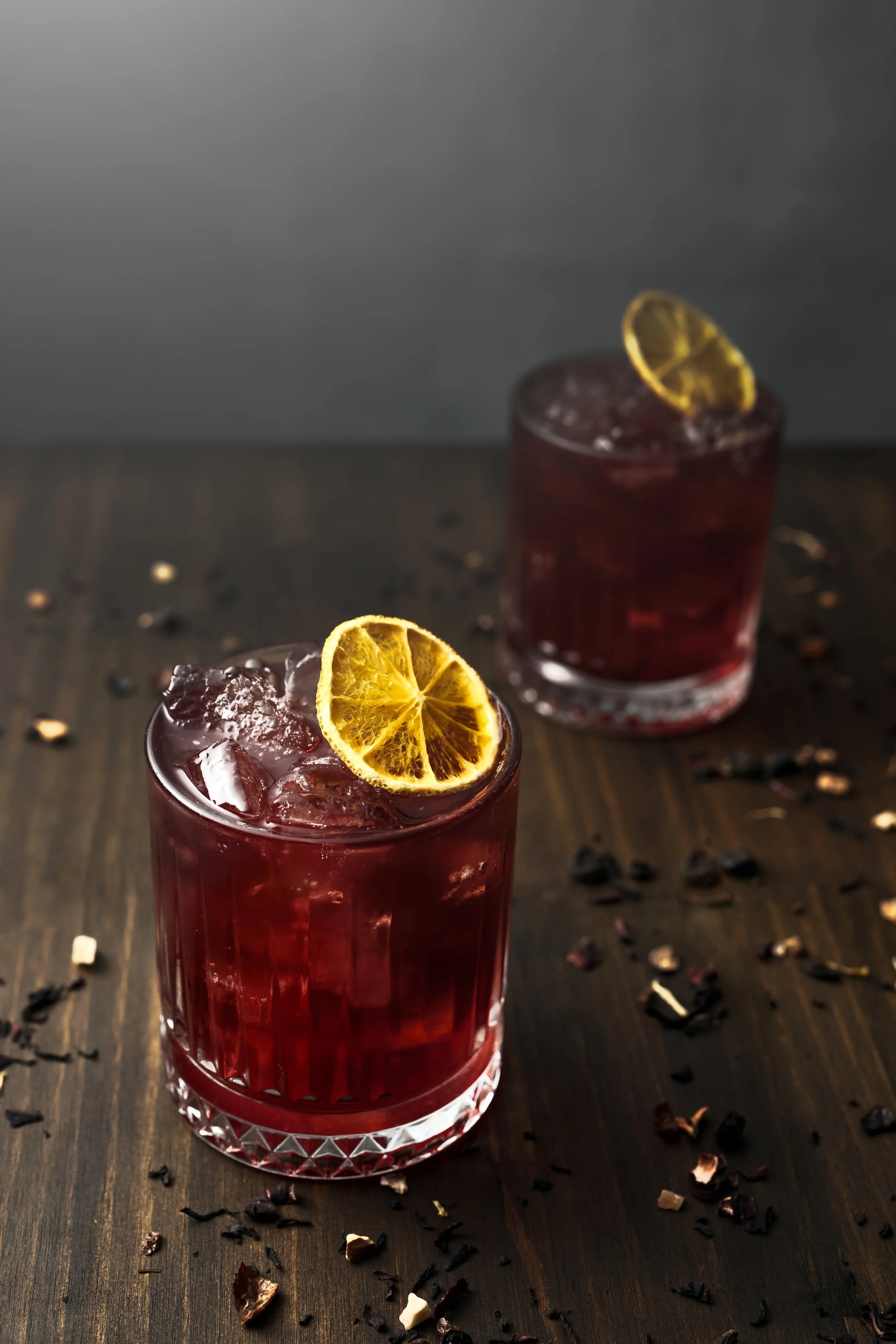 Two glasses of dark red beverage garnished with dried orange slices, on a wooden surface with scattered dried fruit and spices.
