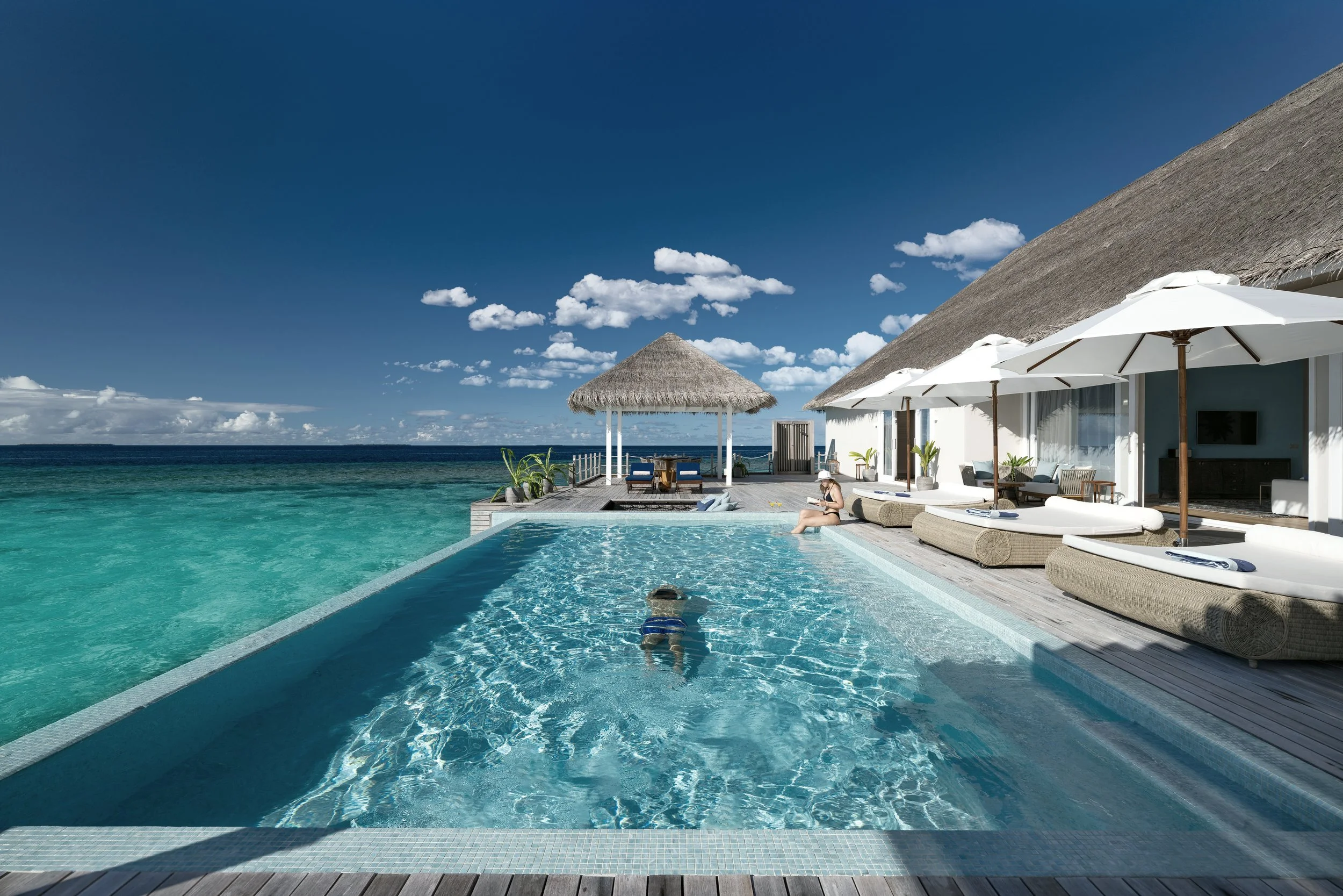 Oceanfront infinity pool with clear water reflecting the blue sky, surrounded by a wooden deck with lounge chairs and umbrellas, overlooking the sea with islands in the distance and a thatched-roof gazebo on the deck.