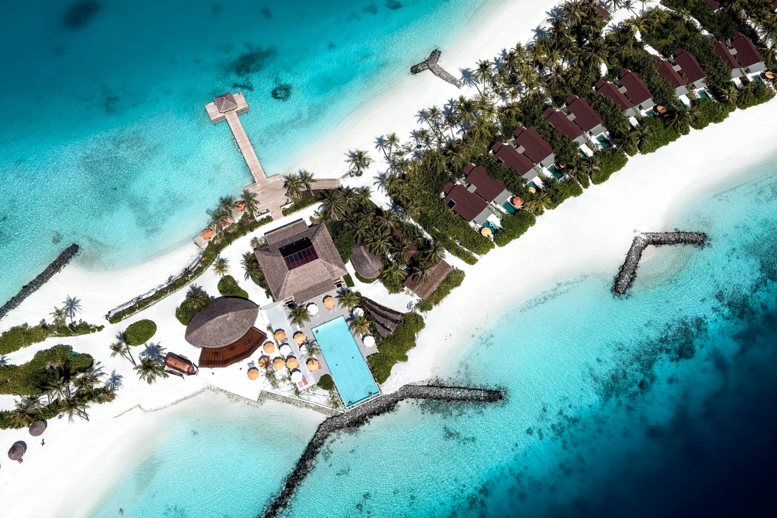 Aerial view of a tropical island resort with overwater bungalows, a swimming pool, palm trees, golfing breakwaters, and turquoise ocean water.