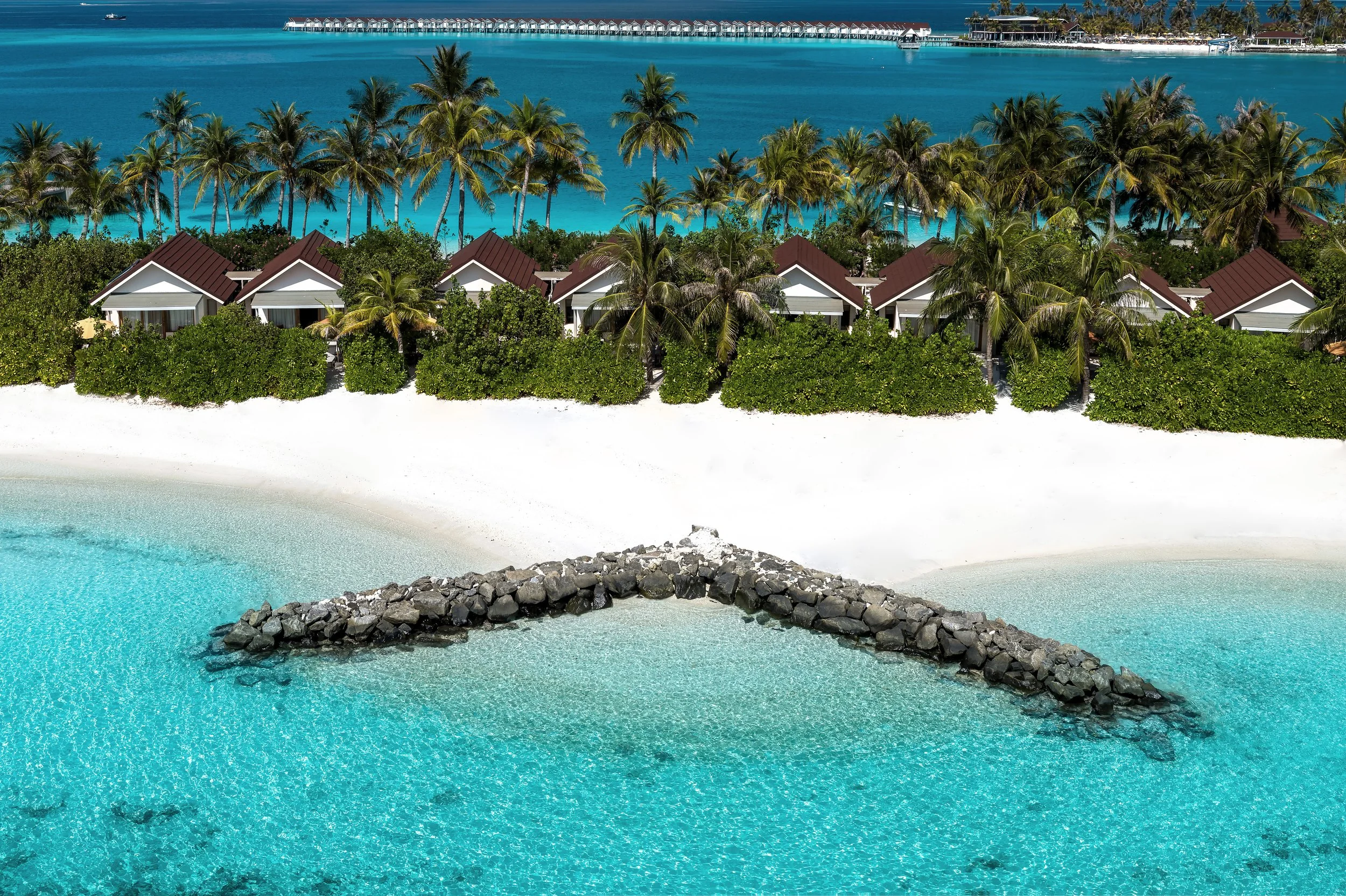 Resort overwater bungalows with a dock, tropical palm trees, white sandy beach, turquoise waters, and a rocky breakwater in the foreground.