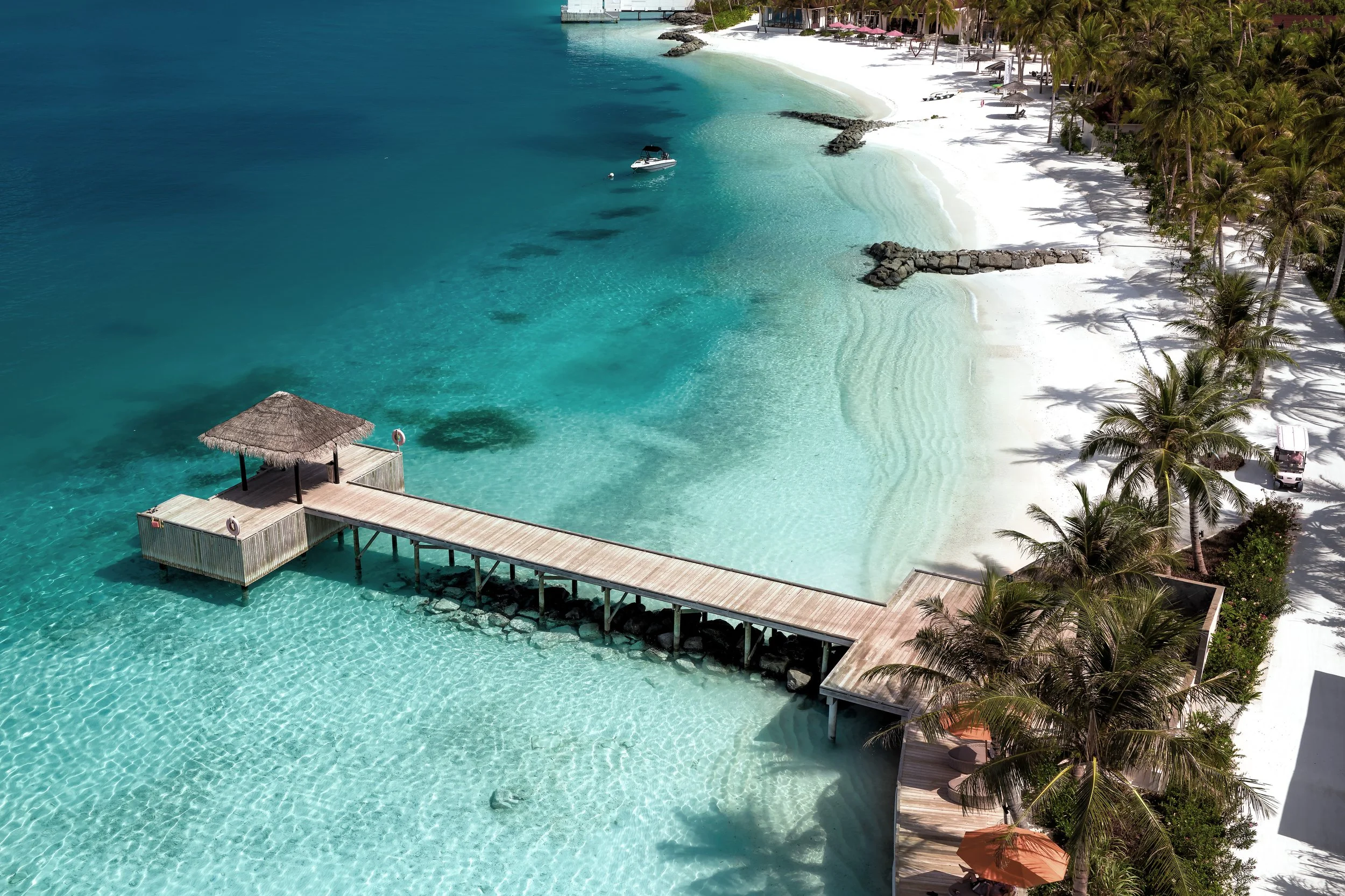 An aerial view of a tropical beach with white sand, turquoise water, palm trees, and a wooden pier with a thatched-roof hut extending over the water. There are lounge chairs and umbrellas along the shoreline, with buildings and greenery in the backgr