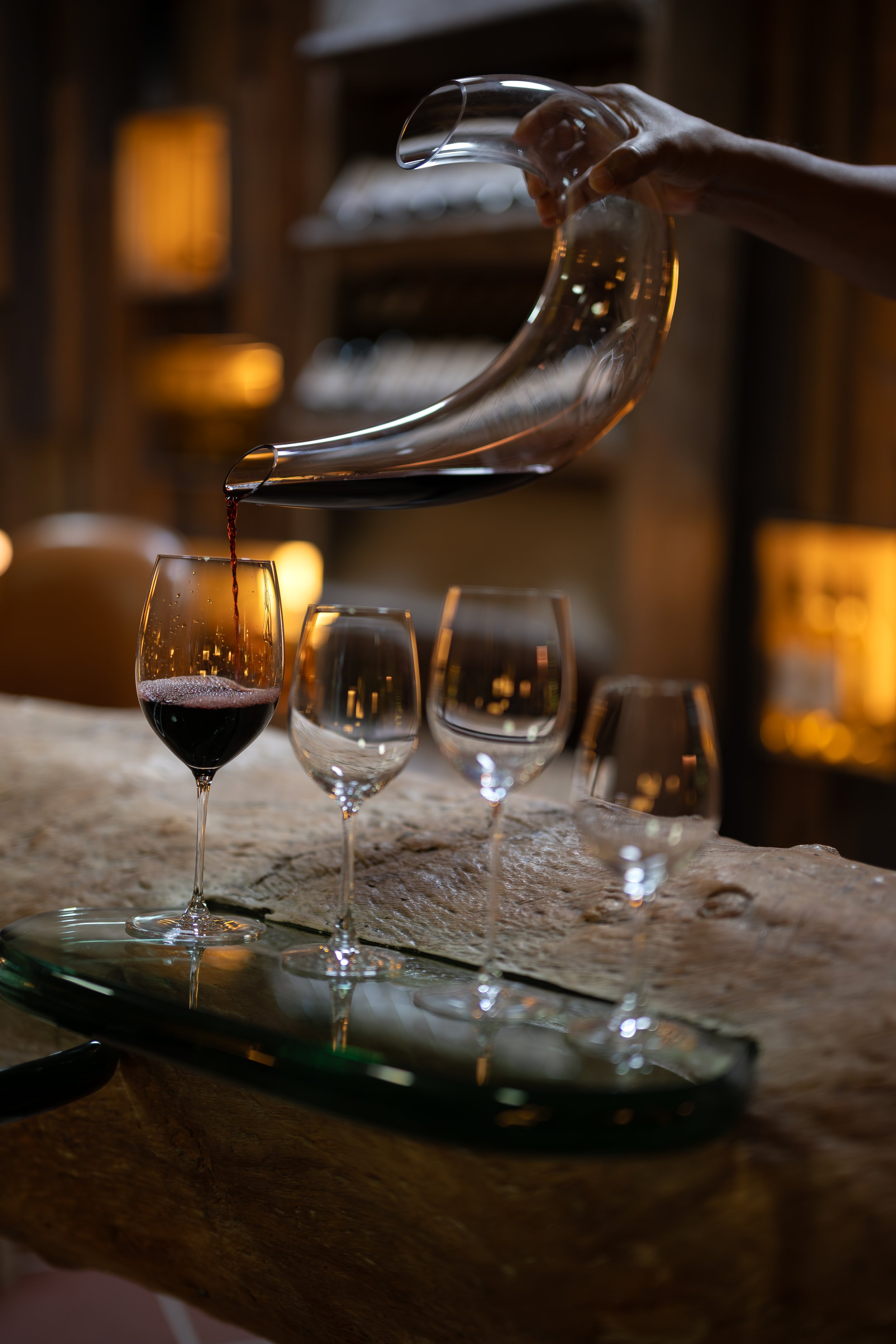 A hand pouring red wine from a decanter into a wine glass on a rustic wooden table, with more empty wine glasses nearby and warm ambient lighting in the background.