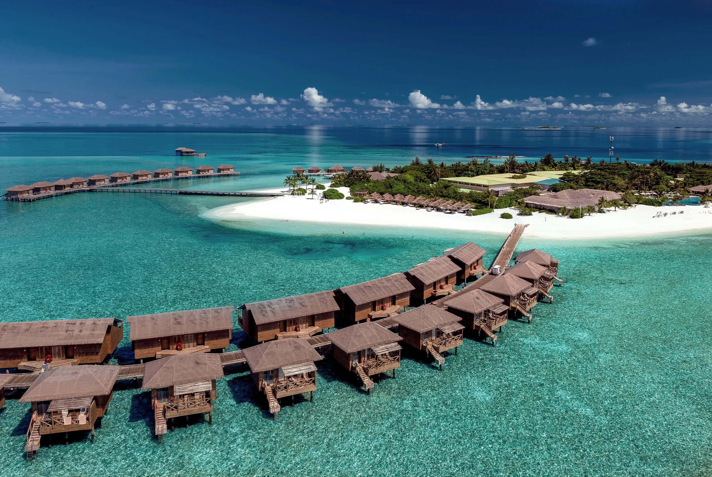 Overwater bungalows extending over turquoise ocean waters near a sandy beach with lush greenery, under a blue sky with scattered clouds.