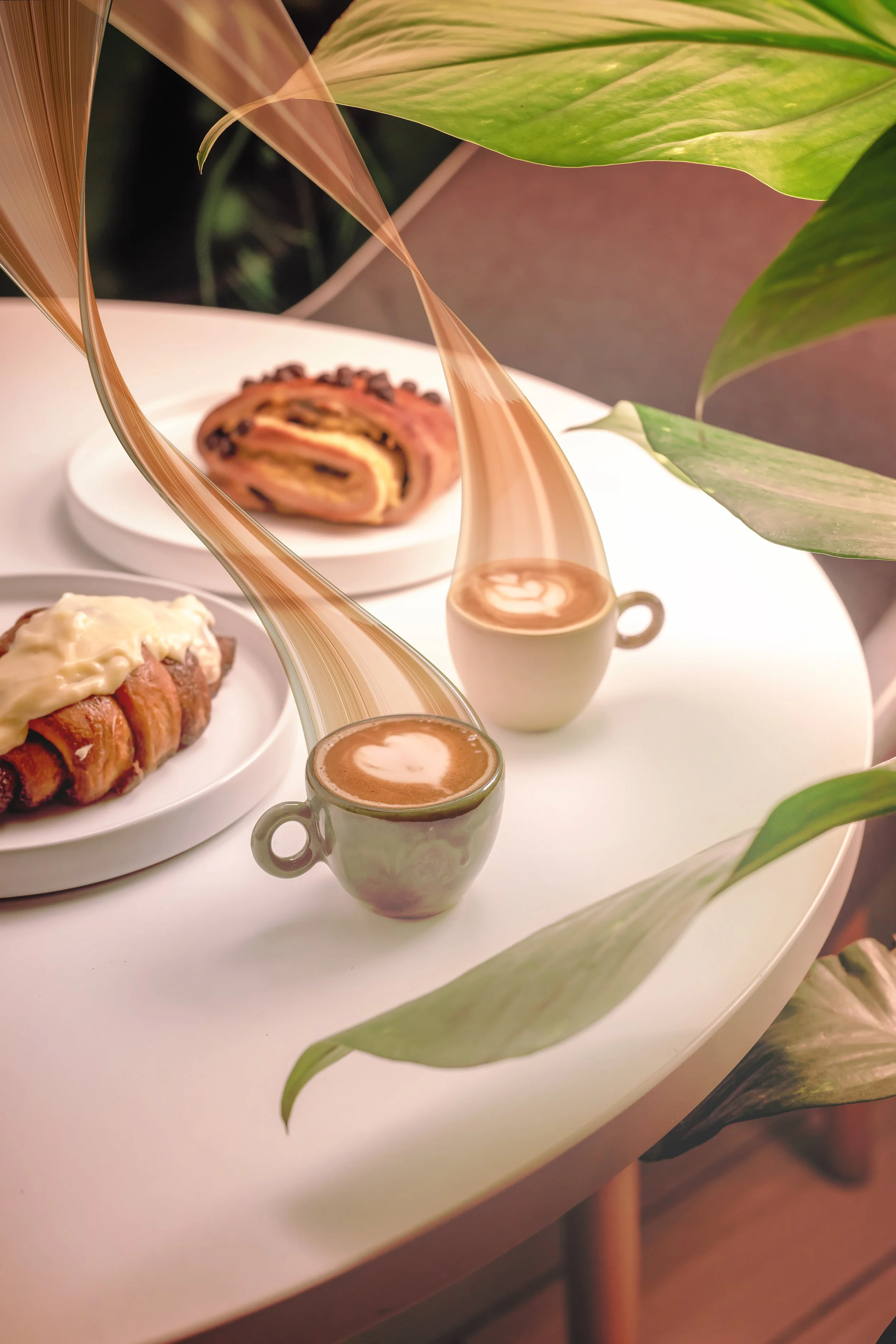 Multiple desserts including a chocolate roll cake on a white plate, a pastry topped with icing, and two cups of coffee on a white table surrounded by green leaves.