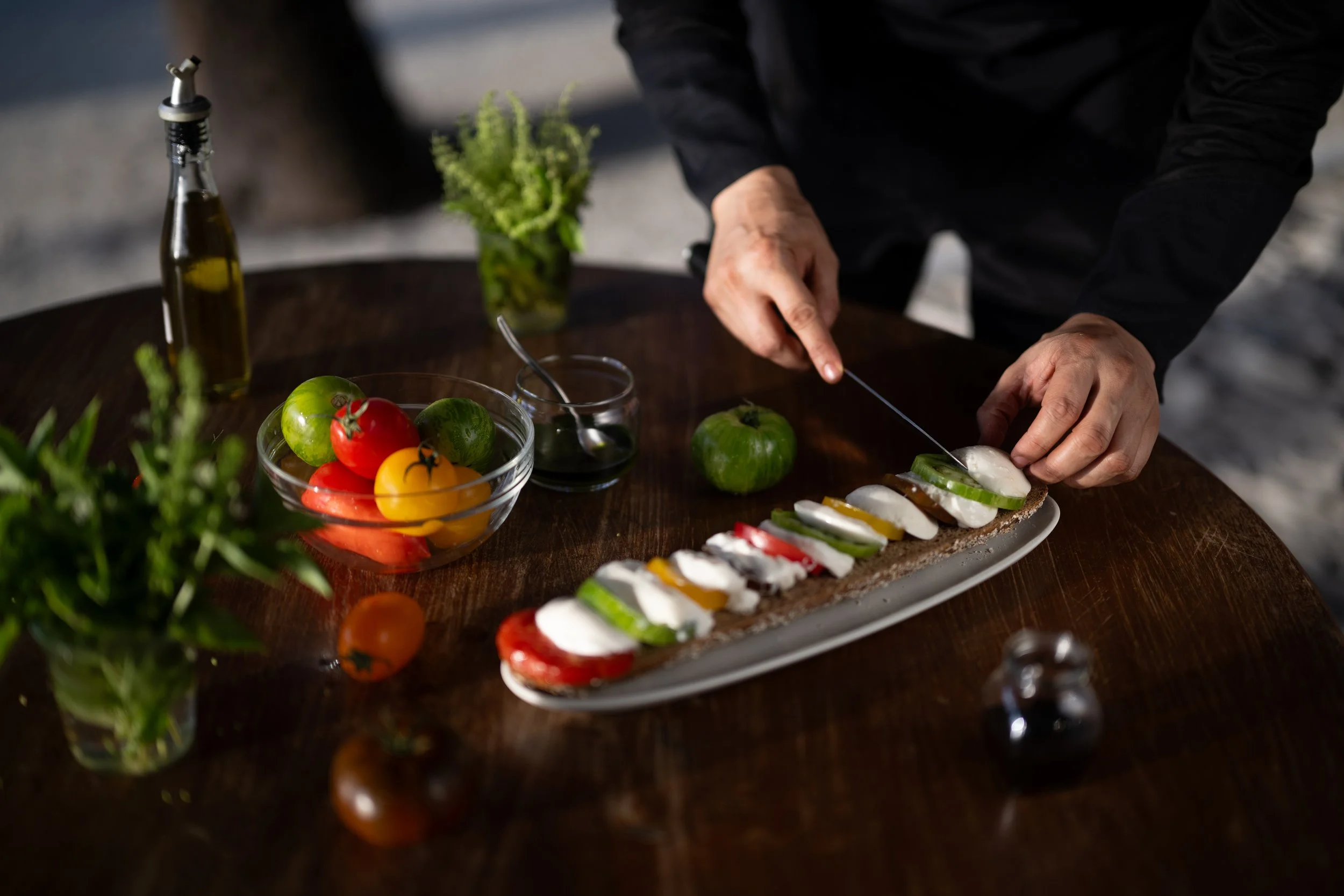 A person preparing an appetizer or snack with sliced vegetables on a rectangular plate, surrounded by fresh tomatoes, limes, herbs, and condiments, on a wooden table.