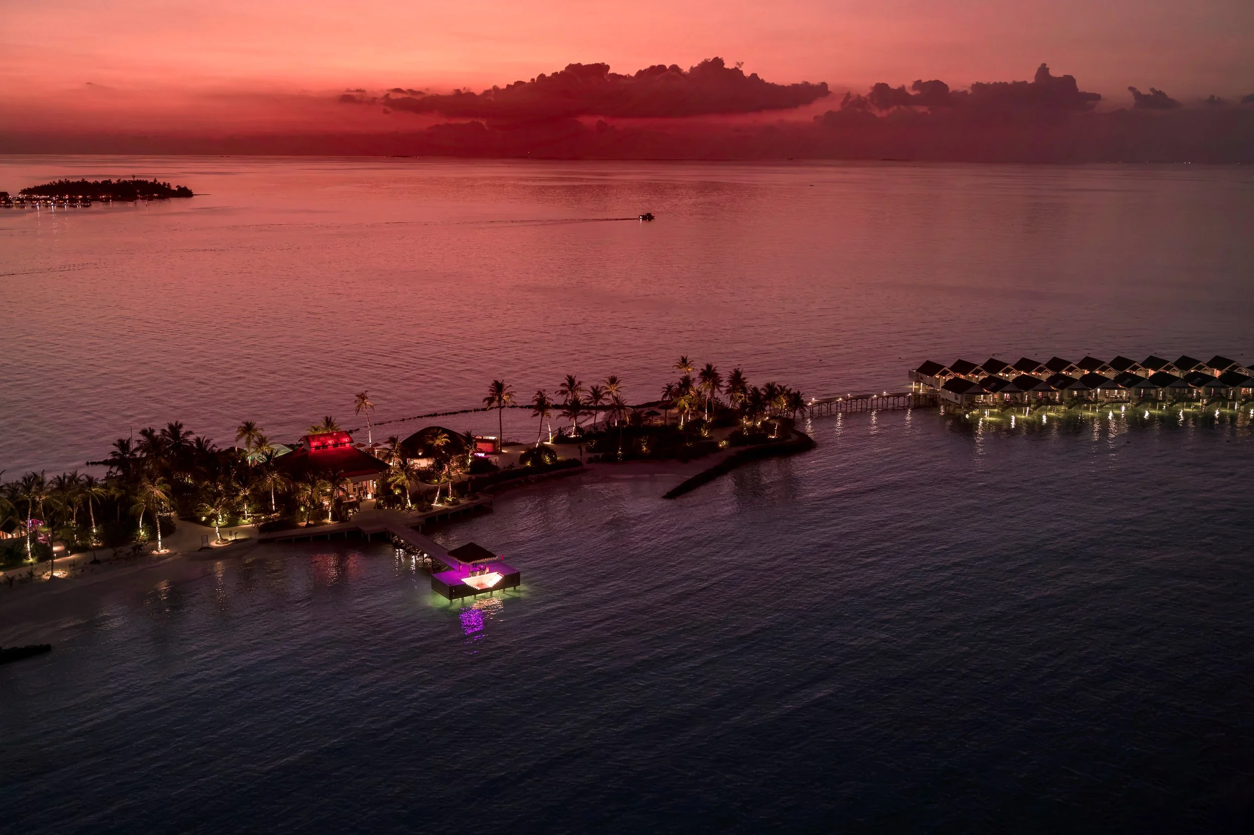 A pink and purple sunset over a calm body of water with overwater bungalows and palm trees along the shoreline.