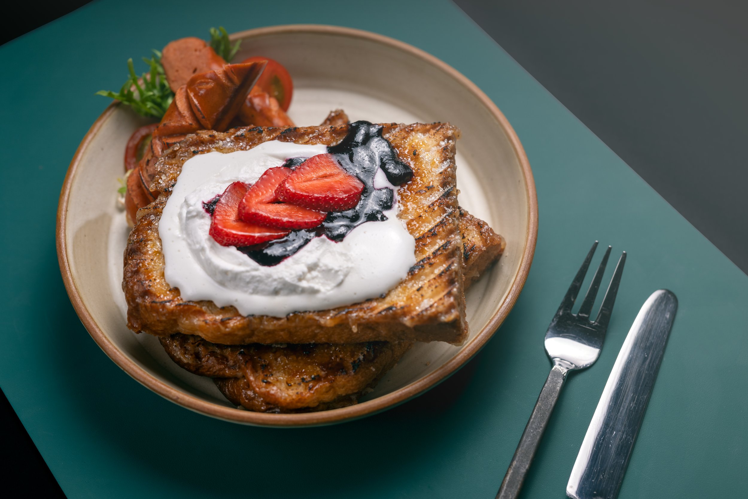A bowl of French toast topped with strawberries, whipped cream, and blueberry syrup, with a side of cherry tomatoes and greens.