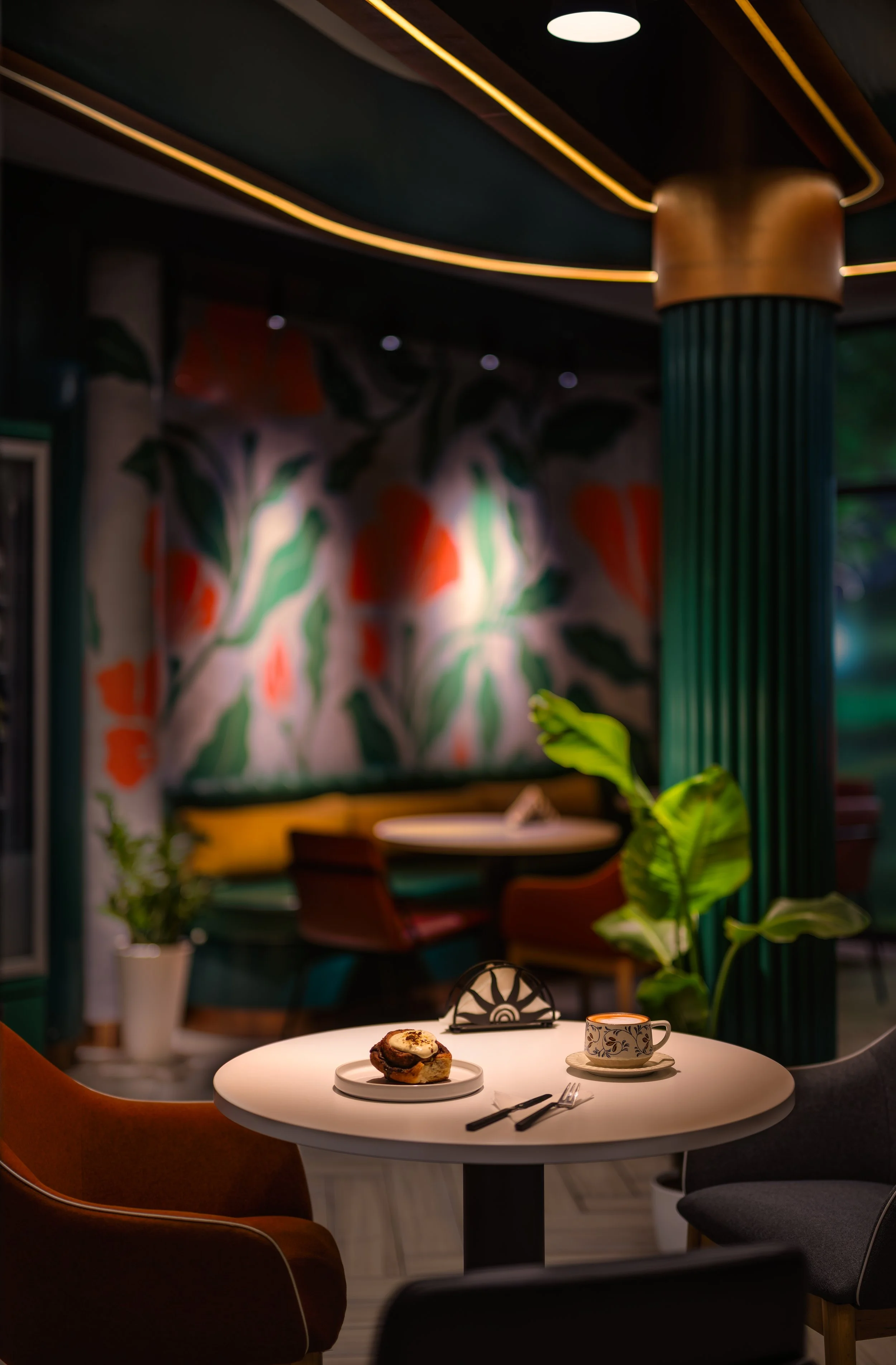 A cozy indoor scene with a round white table set with a cinnamon roll on a white plate, a coffee cup with a floral pattern, a spoon, and black-handled cutlery. Behind the table, there are colorful chairs, green plants, and a mural with orange and gre