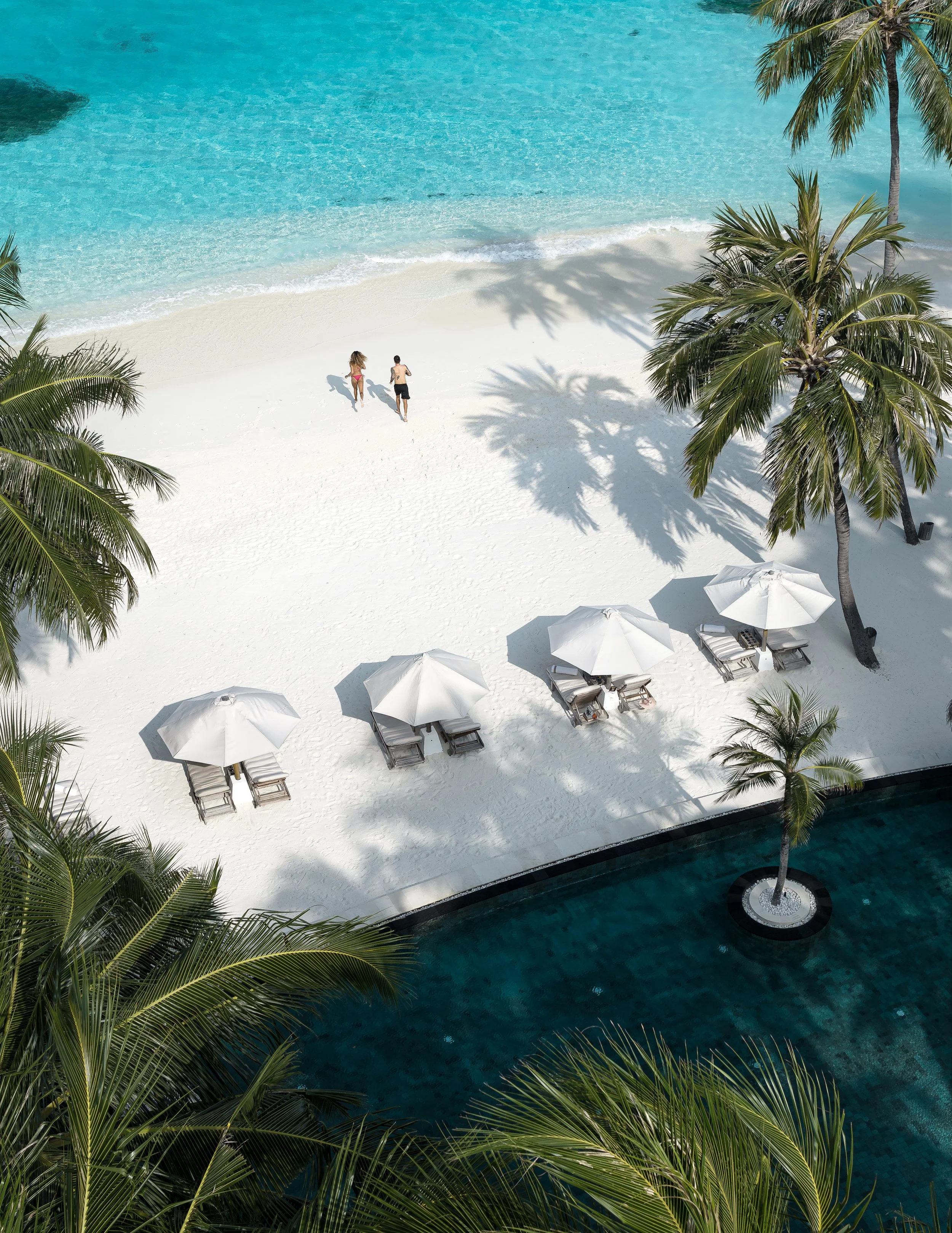 Aerial view of a tropical beach with turquoise water, white sand, palm trees, and lounge chairs with white umbrellas by the poolside. Two people are walking along the shore.