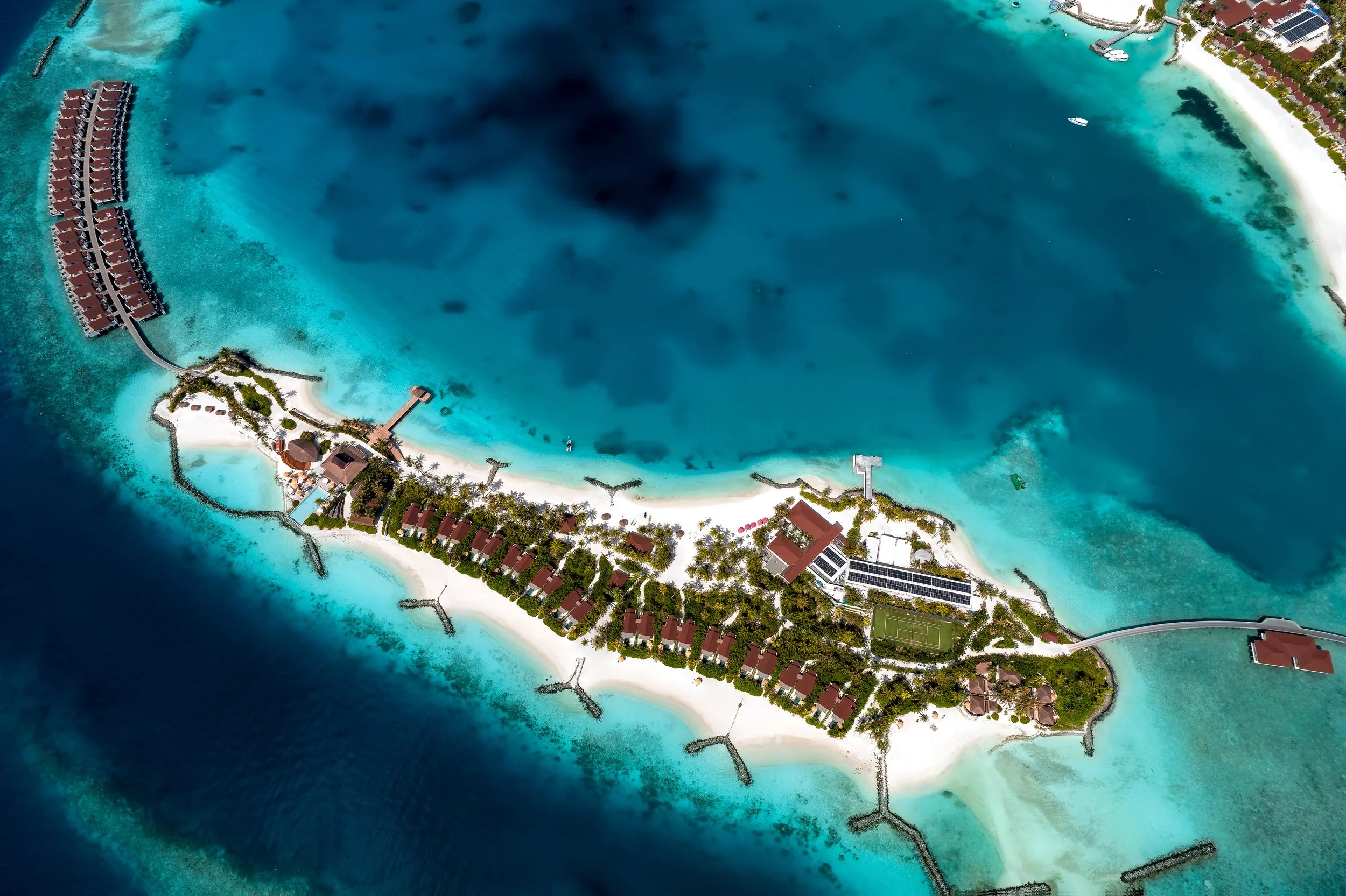 An aerial view of a tropical island resort with overwater bungalows, a sandy beach, and turquoise waters, surrounded by a barrier reef.