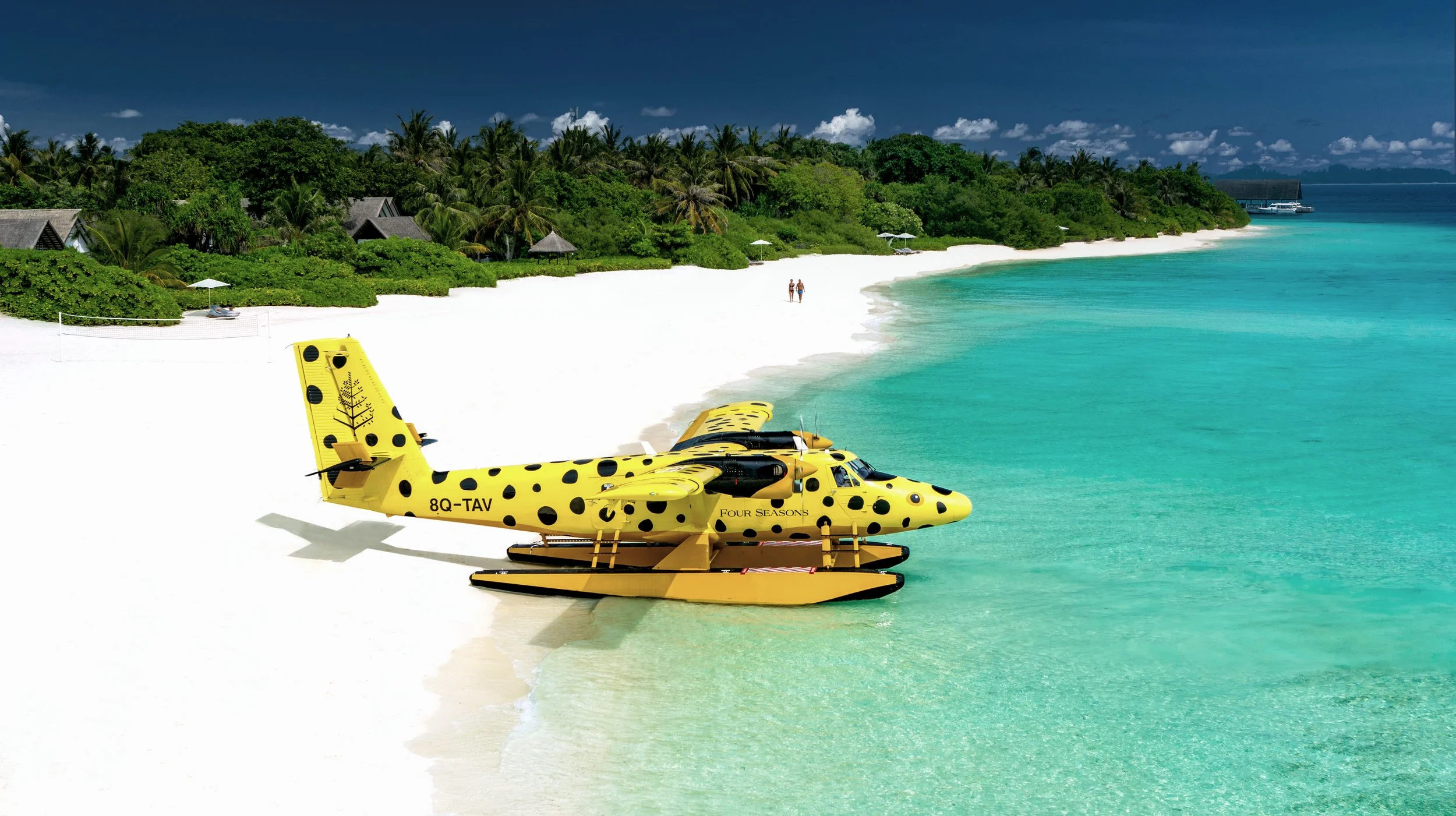 Yellow amphibious aircraft with black spots parked on a white sandy beach near clear turquoise water, with green lush trees and huts in the background.