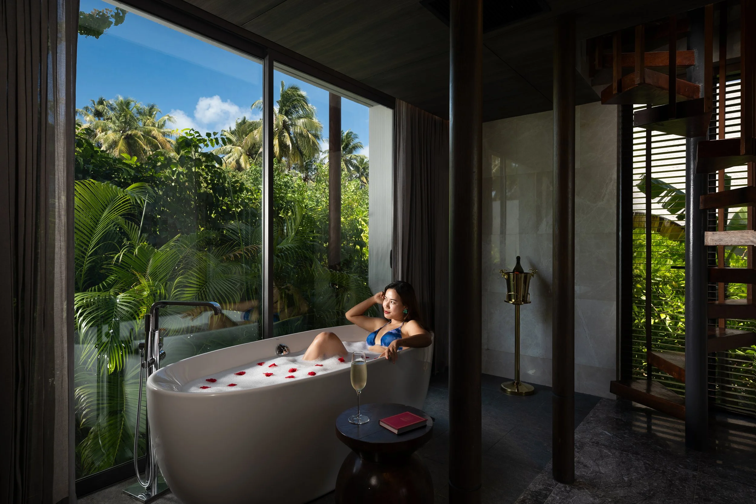 A woman relaxing in a bathtub filled with bubbles and rose petals, holding a glass of wine in a luxurious bathroom surrounded by large windows with views of lush tropical trees and blue sky.