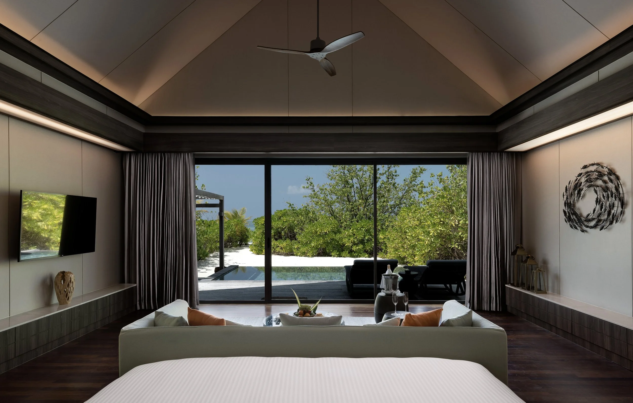 Modern bedroom with large sliding glass doors opening to outdoor greenery, decorated with a wall-mounted flat-screen TV, vases, and wall art, featuring a coffered ceiling with a ceiling fan.
