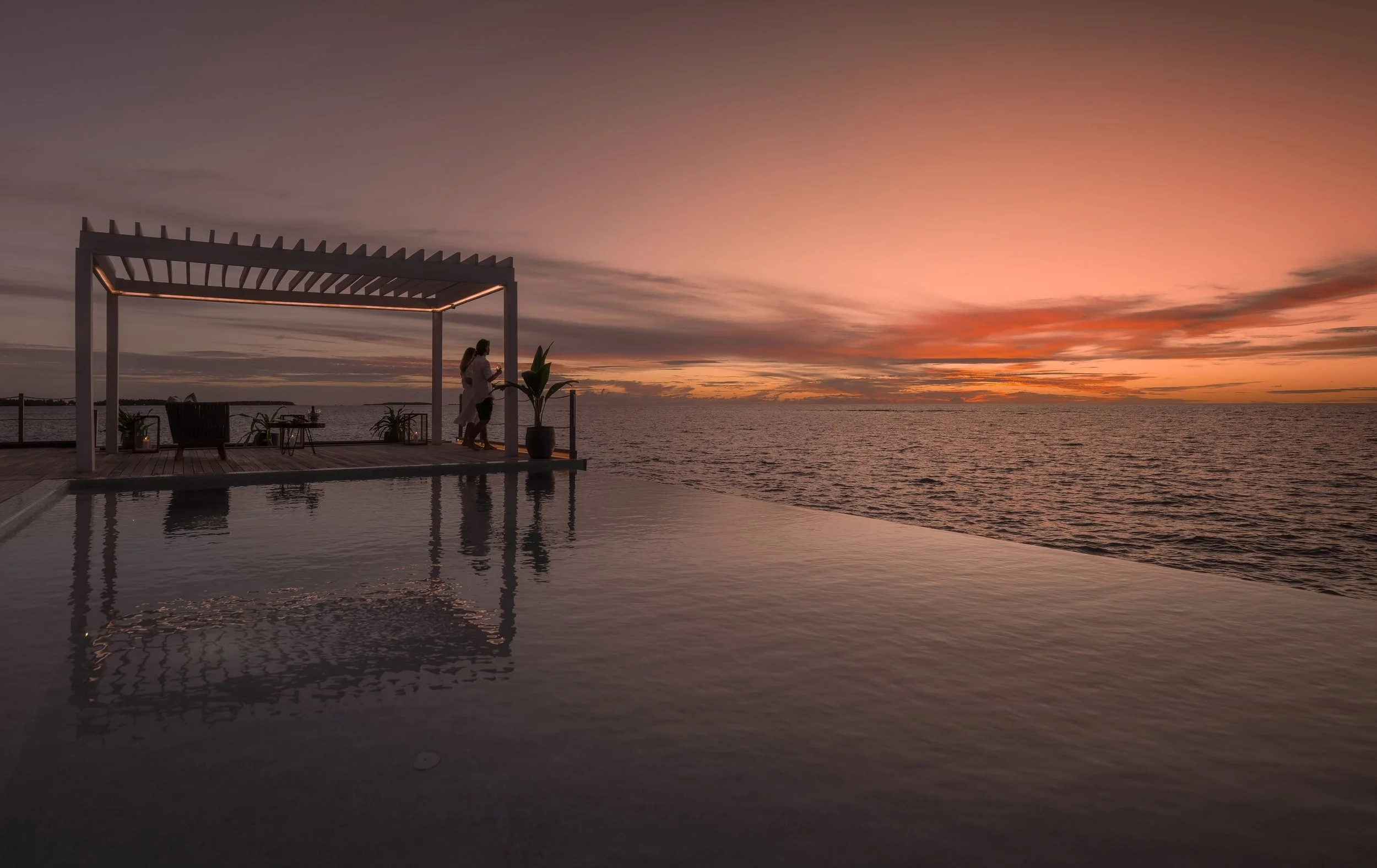 Couple standing on a deck overlooking the ocean at sunset with a pergola structure overhead and potted plants nearby.