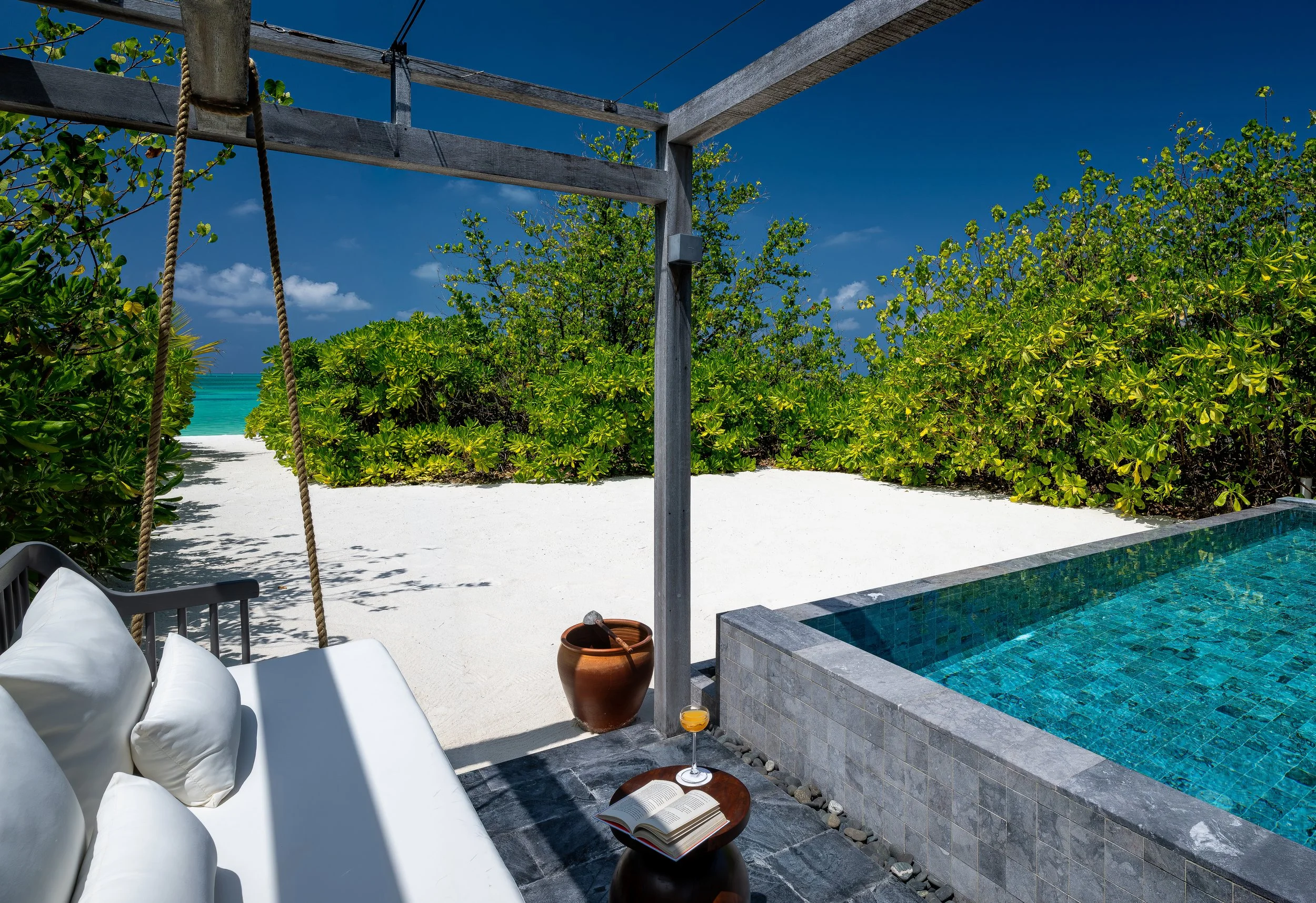Outdoor rooftop lounge area with white cushioned seating, a swing with rope, a small round table with an open book and a glass of orange juice, a terracotta pot, a hot tub with blue tiles, lush green bushes, and a view of the ocean in the background 