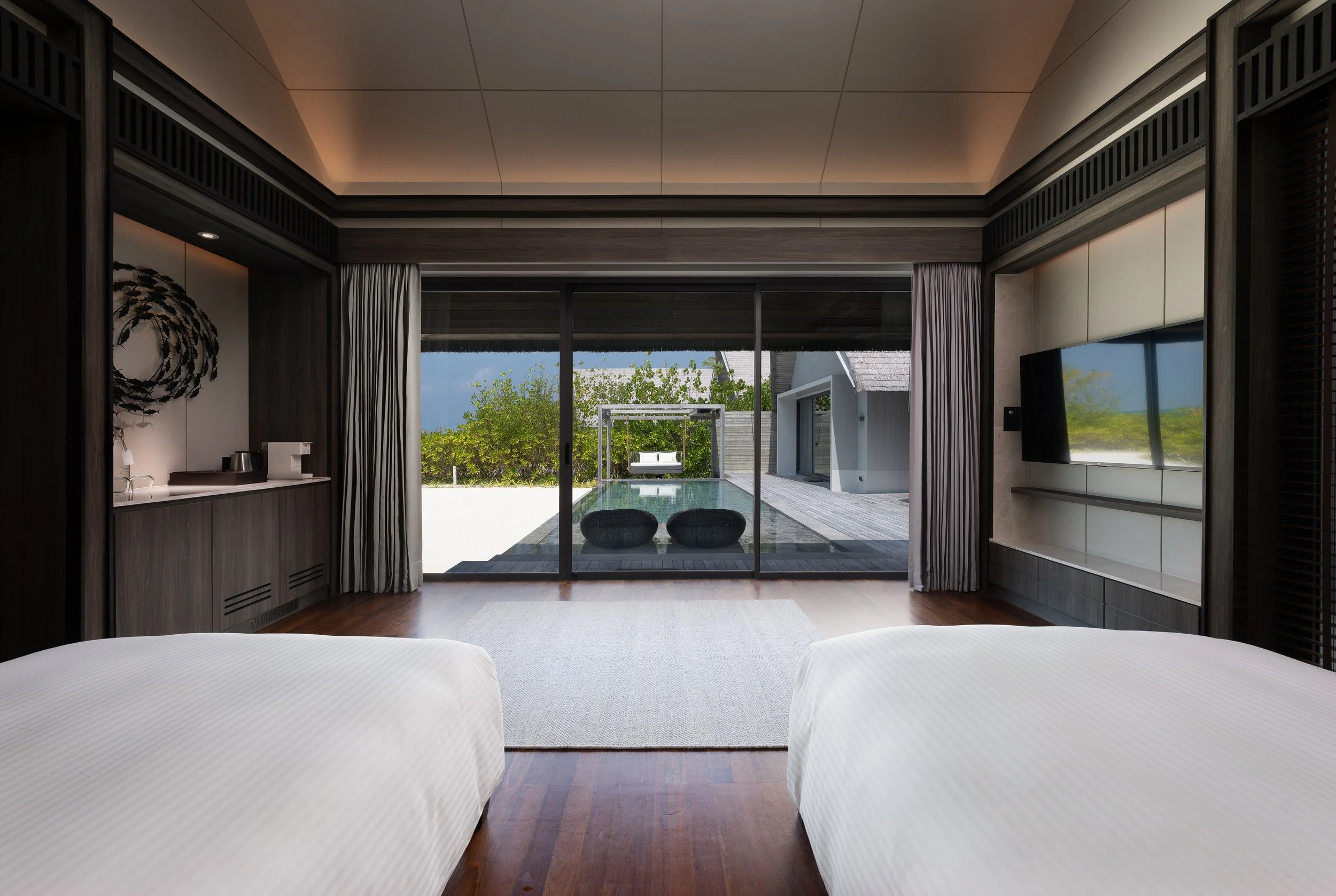 The image depicts the interior of a modern bedroom with two beds, a large glass sliding door leading to an outdoor patio with a pool, and lush greenery outside. The room features dark wood accents, light-colored curtains, and a minimalist design.