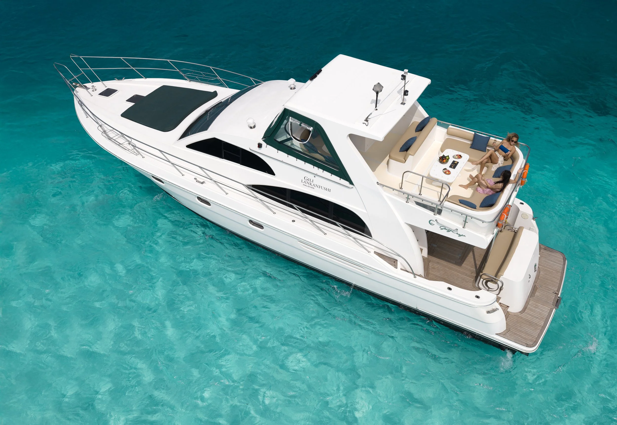 A white yacht anchored in turquoise water with two women relaxing on the deck, one reading a book and the other sitting near a small table with snacks.