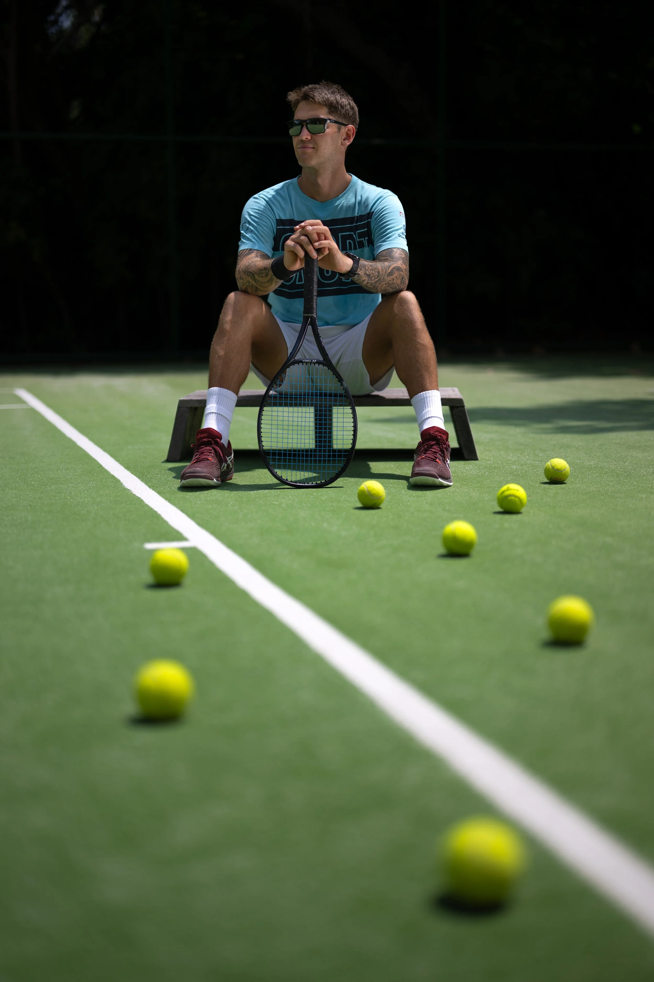 A man sitting on a bench on a tennis court, with tennis balls scattered around him. He is wearing sunglasses, a light blue shirt, white shorts, and red shoes, holding a tennis racket, and looking into the distance.
