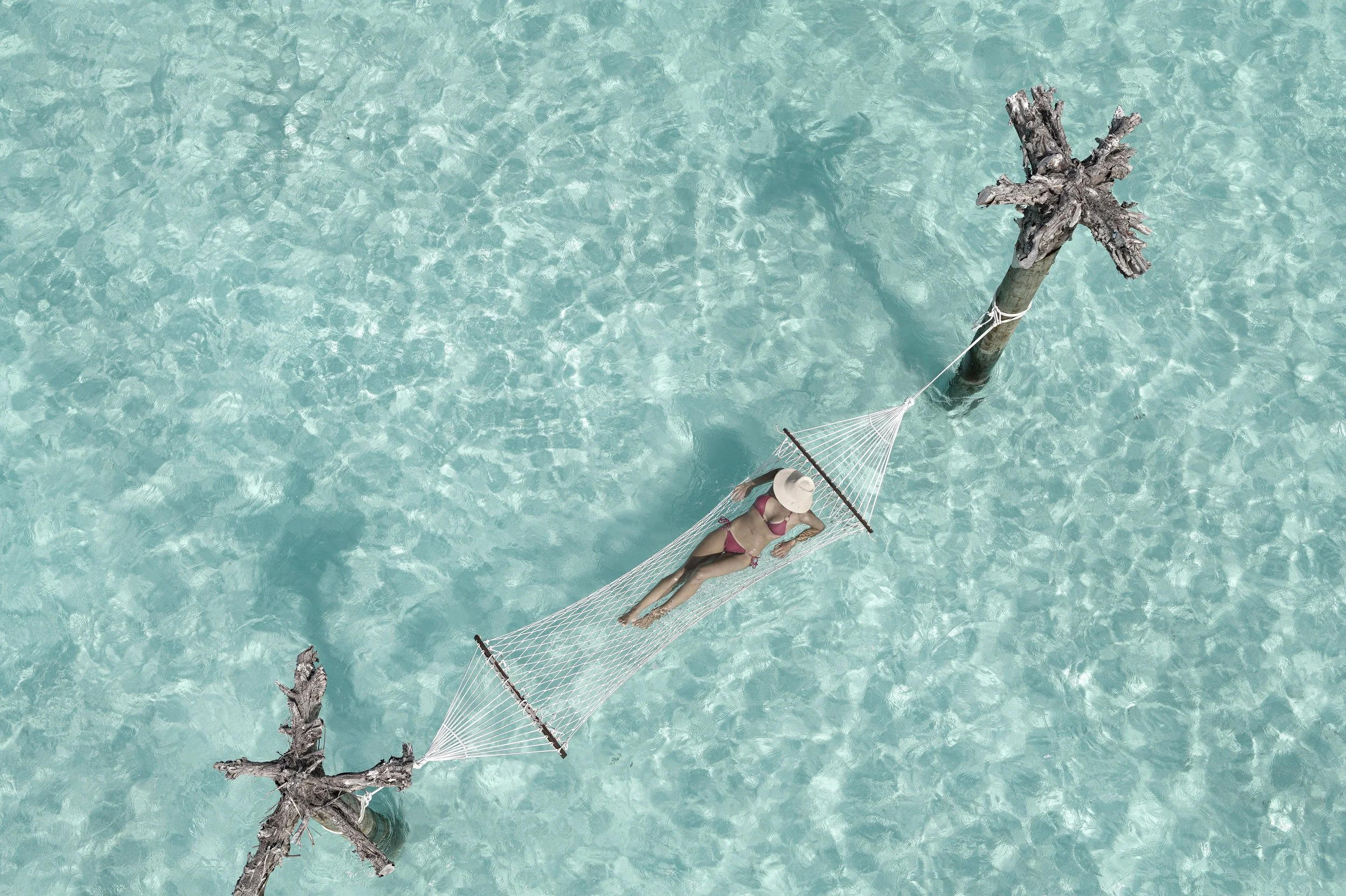 A woman in a pink bikini and wide-brim hat relaxing on a hammock tied between two weathered trees over clear turquoise water.