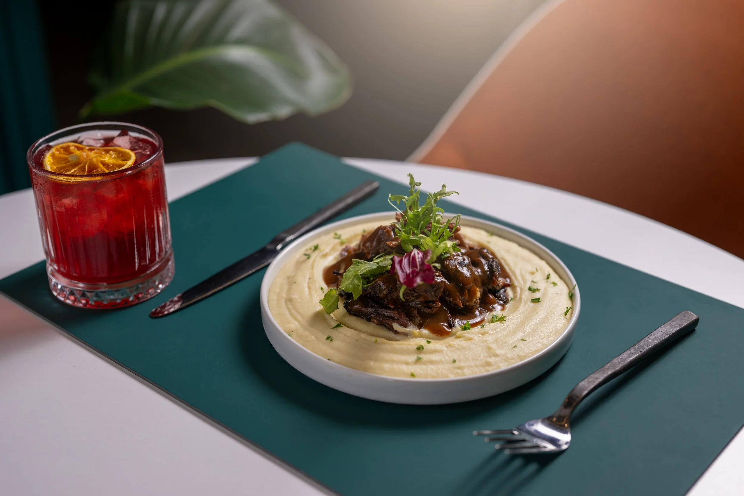 A bowl of mashed potatoes topped with beef stew and garnished with greens. A glass of red drink with a lemon slice sits beside it on a teal tray, with a fork and knife nearby on a white table.