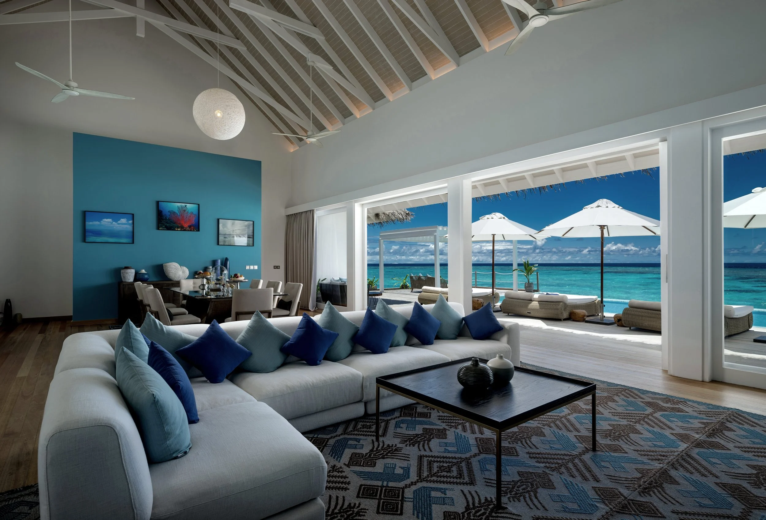 Bright living room with white sofa and blue pillows, black coffee table, patterned rug, and ocean view through large open windows with white umbrellas on patio.