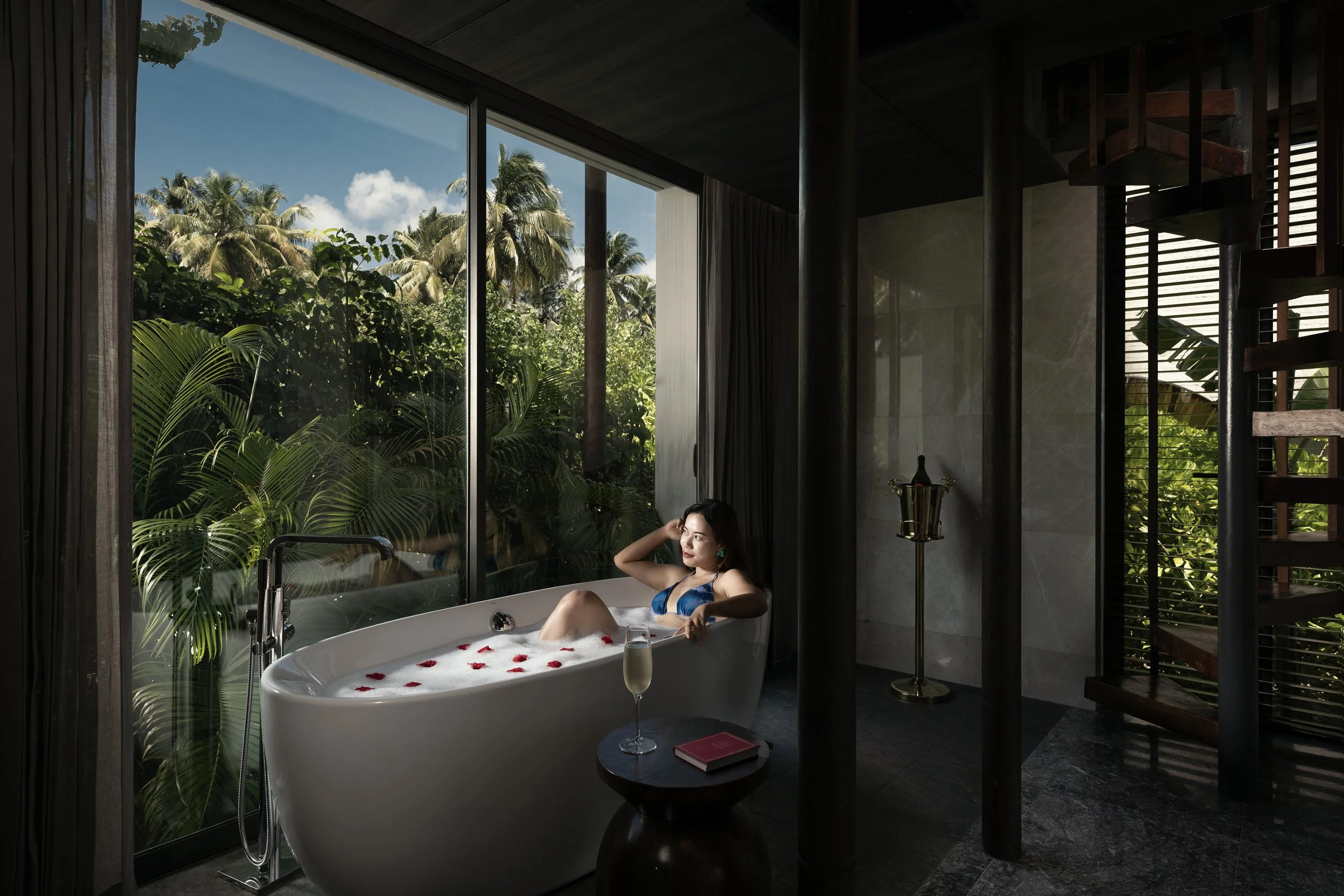 A woman relaxing in a bathtub filled with bubbles and rose petals, with a glass of champagne and a book nearby, inside a modern bathroom with large windows revealing lush green tropical trees outside.