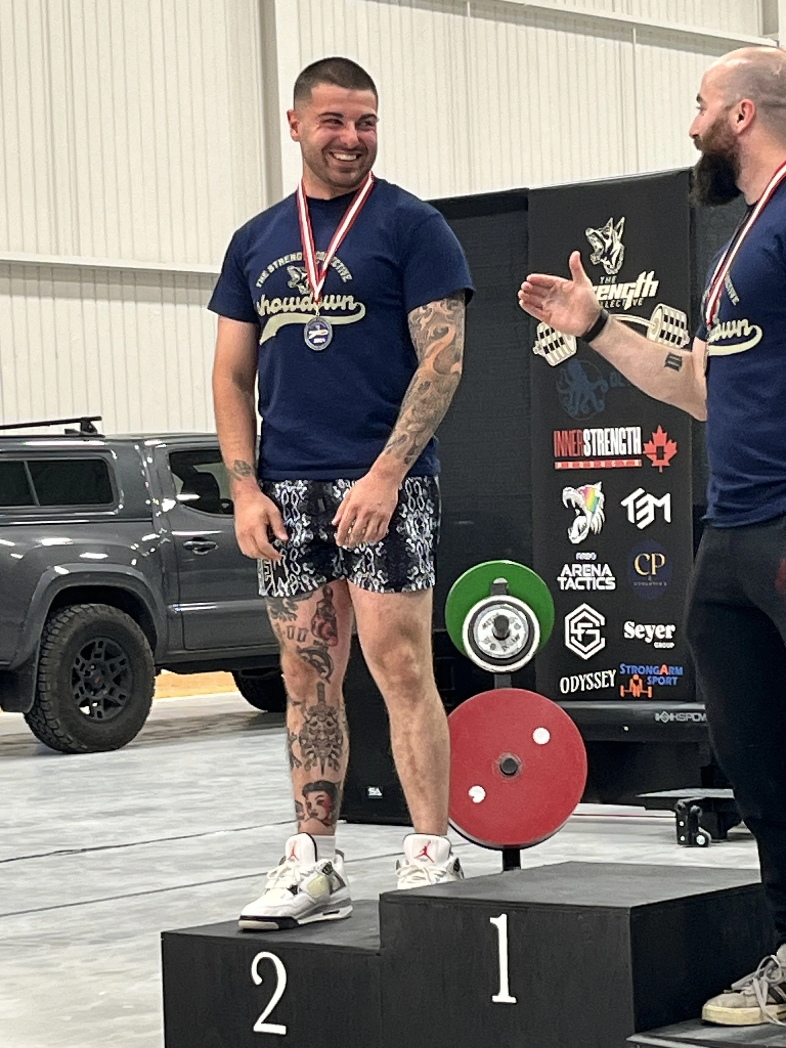 A man standing on a winners' podium with a medal around his neck, smiling at another man who is talking to him. The man on the podium is wearing a navy t-shirt, patterned shorts, and white sneakers. The background shows a black sign with various logos and a green and red weight plate.