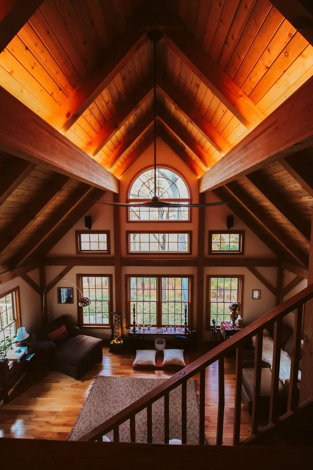 Interior view of a cabin or loft with a high, wooden, vaulted ceiling and large windows, including a prominent arched window. The room is furnished with a dark sofa, a lamp, cushions, a small table, and decorative items, with warm lighting highlighting the wood paneling and flooring.
