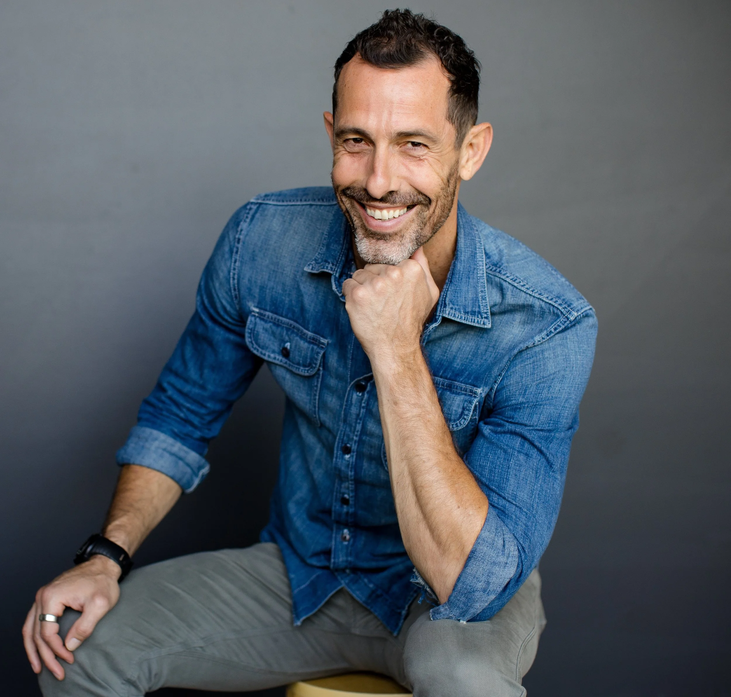 A man with dark hair and a beard, smiling and winking, wearing a blue denim shirt and gray pants, seated against a plain gray background.