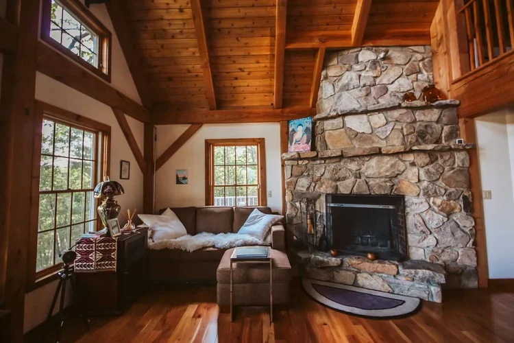 Living room with a large stone fireplace, wooden ceiling, and multiple windows, featuring a brown sectional sofa with white pillows and a furry throw, a small side table, and decorative items on the mantel.
