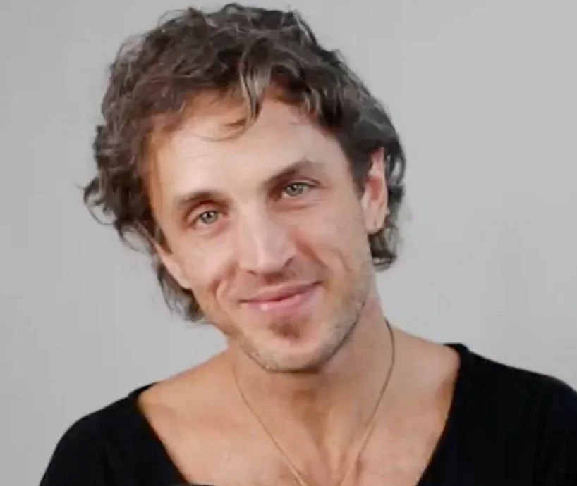 Close-up of a smiling man with light skin, curly hair, and facial hair, wearing a black shirt against a plain background.