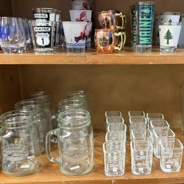 A wooden shelf displaying souvenir Maine-themed glassware, including mugs, cups, and shot glasses, along with etched West Quoddy glasses on a lower shelf.