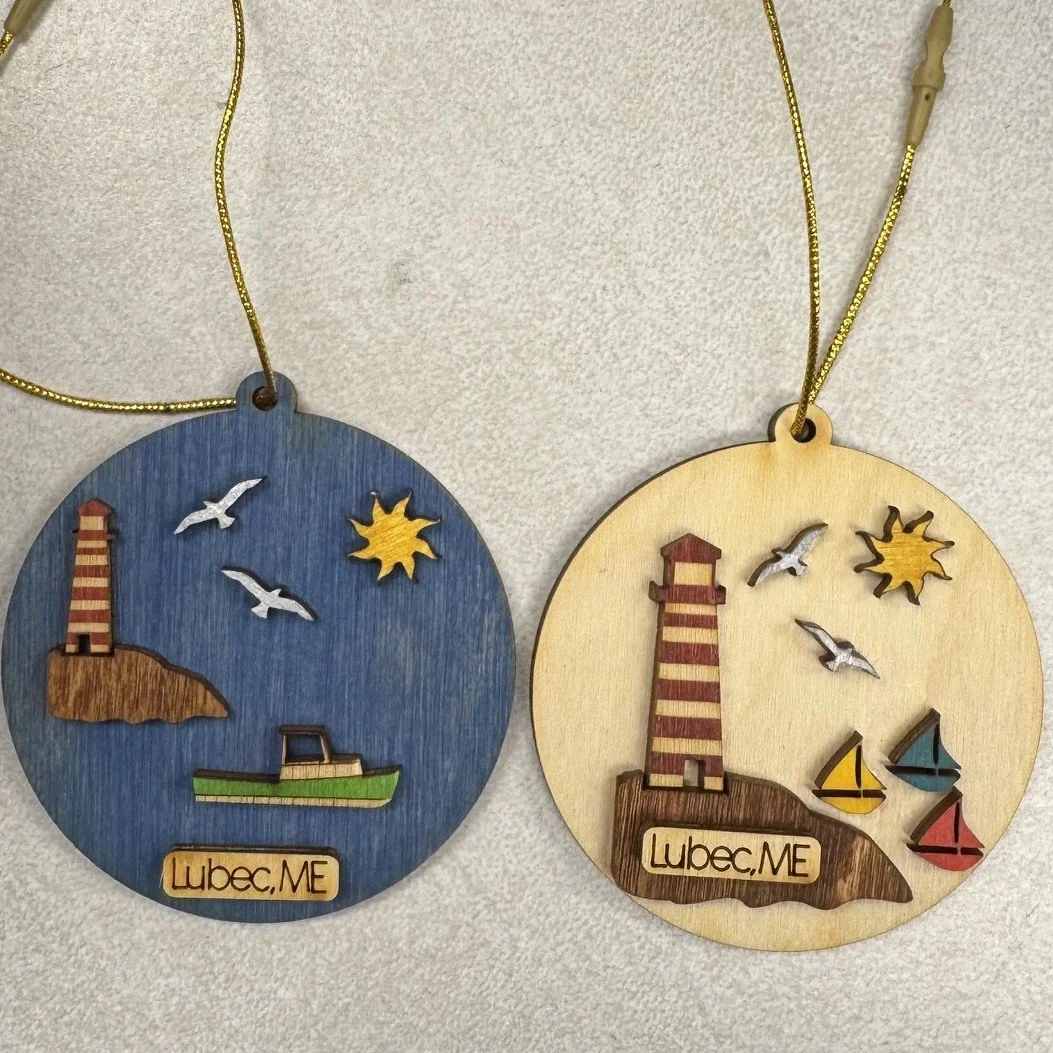 Two round souvenir wooden ornaments with lighthouse, seagulls, and boats, labeled "Lubec, ME"; one is blue with a striped West Quoddy lighthouse, the other is beige with a striped lighthouse and colorful sailboats.