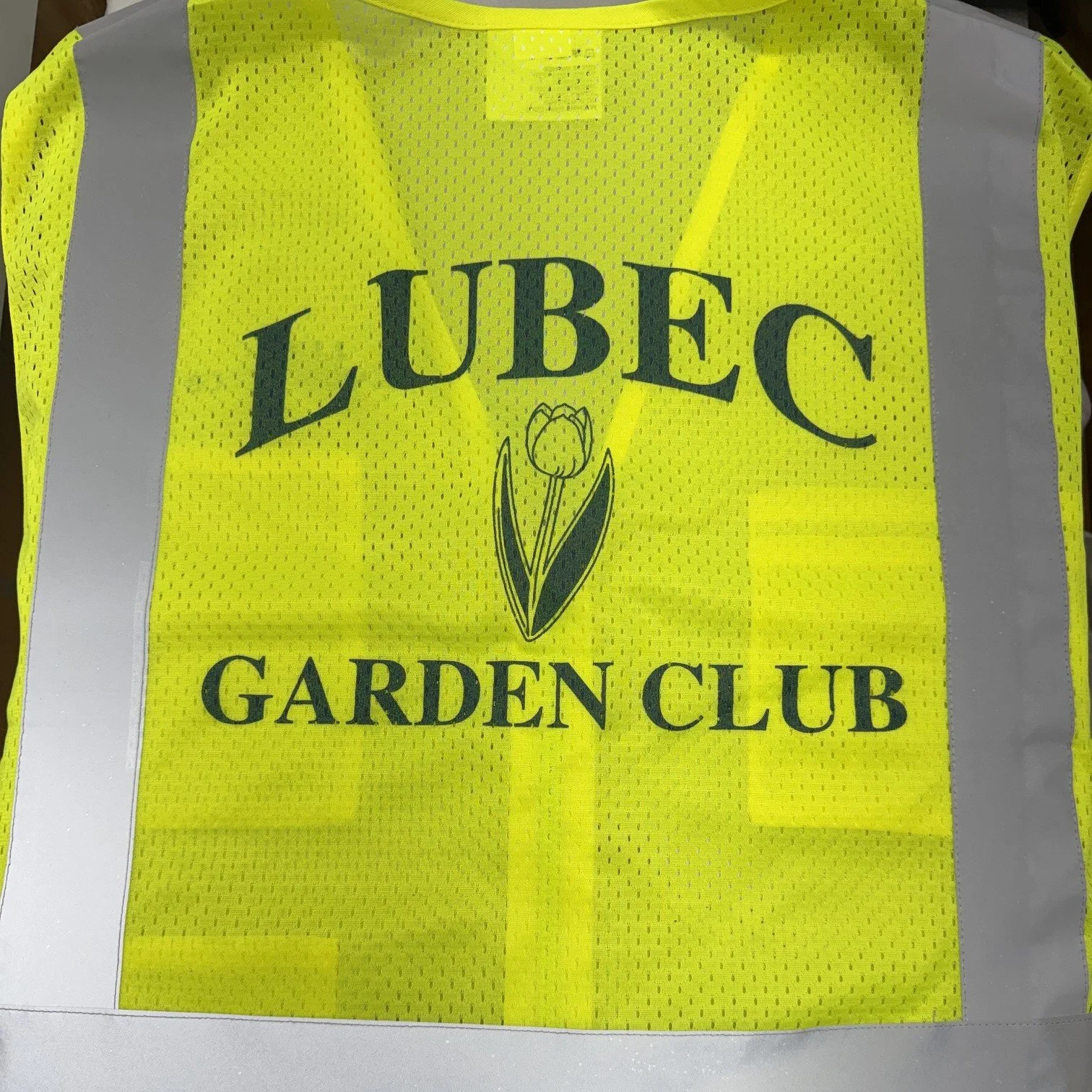 A yellow high-visibility safety vest with reflective silver stripes and the logo of the Lubec Garden Club featuring a tulip flower in the center.