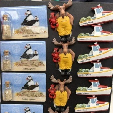 Souvenir Lubec magents including puffins, four moose dressed in yellow overalls with red flowers, and three wooden boats with white hulls and red details arranged vertically.
