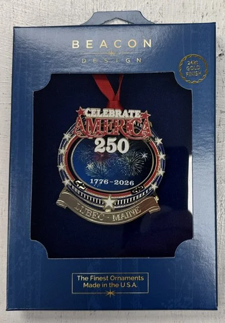 Box with a souvenir ornament celebrating America's 250th anniversary from 1776 to 2026, featuring patriotic fireworks, stars, and the words 'Celebrate America 250' with a ribbon and a banner that says 'LUBEC, MAINE.'
