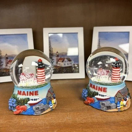 Two souvenir Maine-themed snow globes with lighthouses, seagulls, lobster, and boats, placed on a wooden surface with framed pictures of the lighthouse in the background.