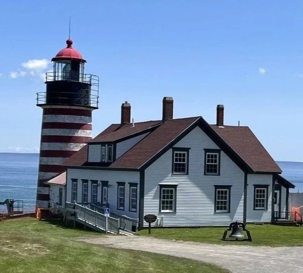 A lighthouse with red and white stripes next to a white house with a brown roof on a grassy area near the ocean.