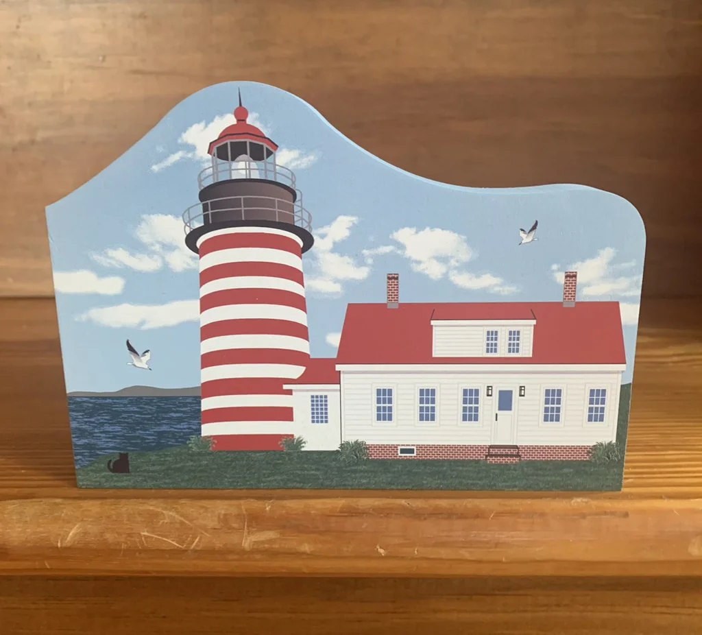 A cartoon illustration of a lighthouse with red and white stripes next to a white house with a red roof, set against a bright blue sky with clouds and two seagulls flying. The scene is on a grassy area by the water.