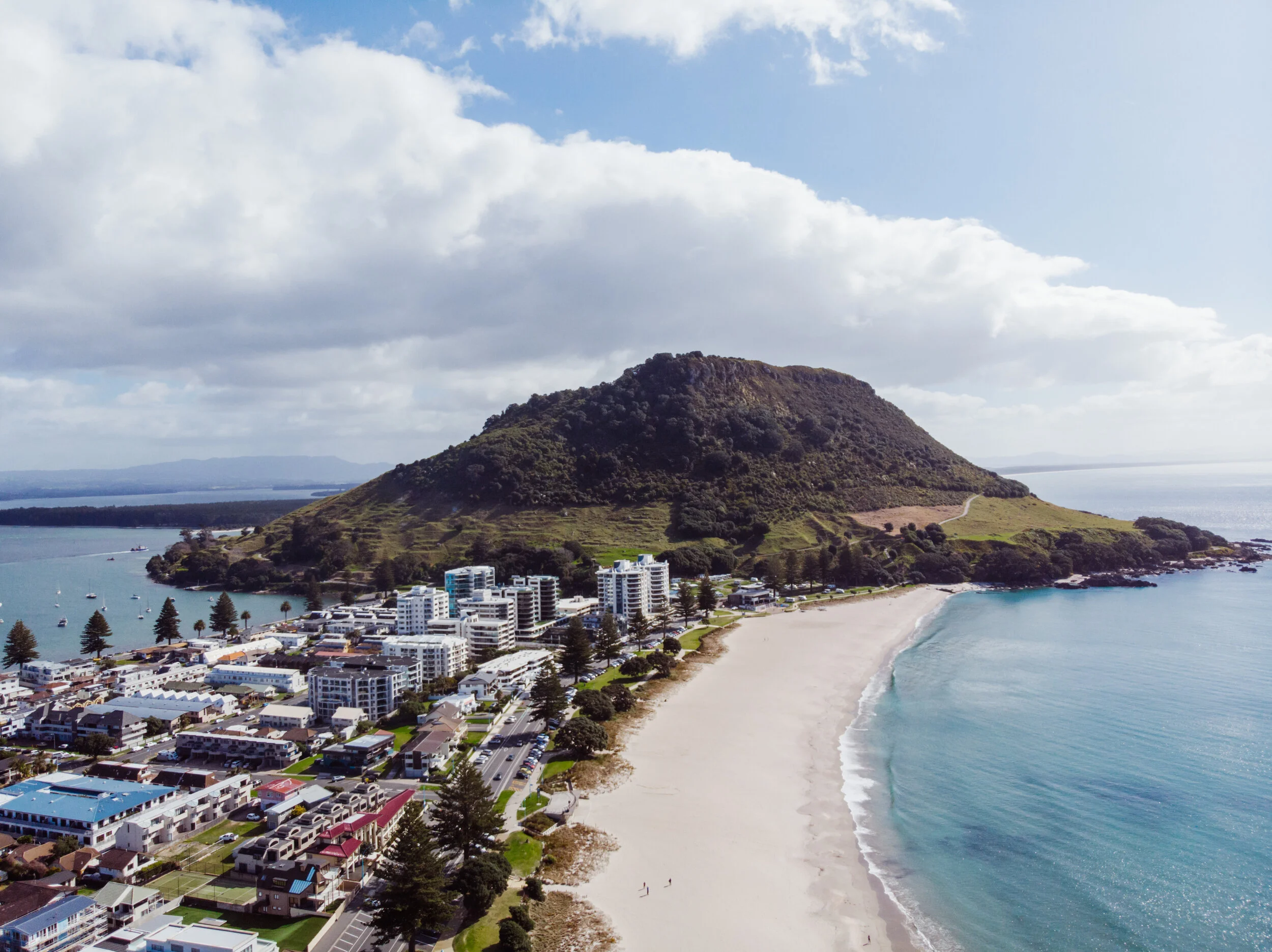A coastal town with modern buildings near a sandy beach and a green hill in the background under a partly cloudy sky.