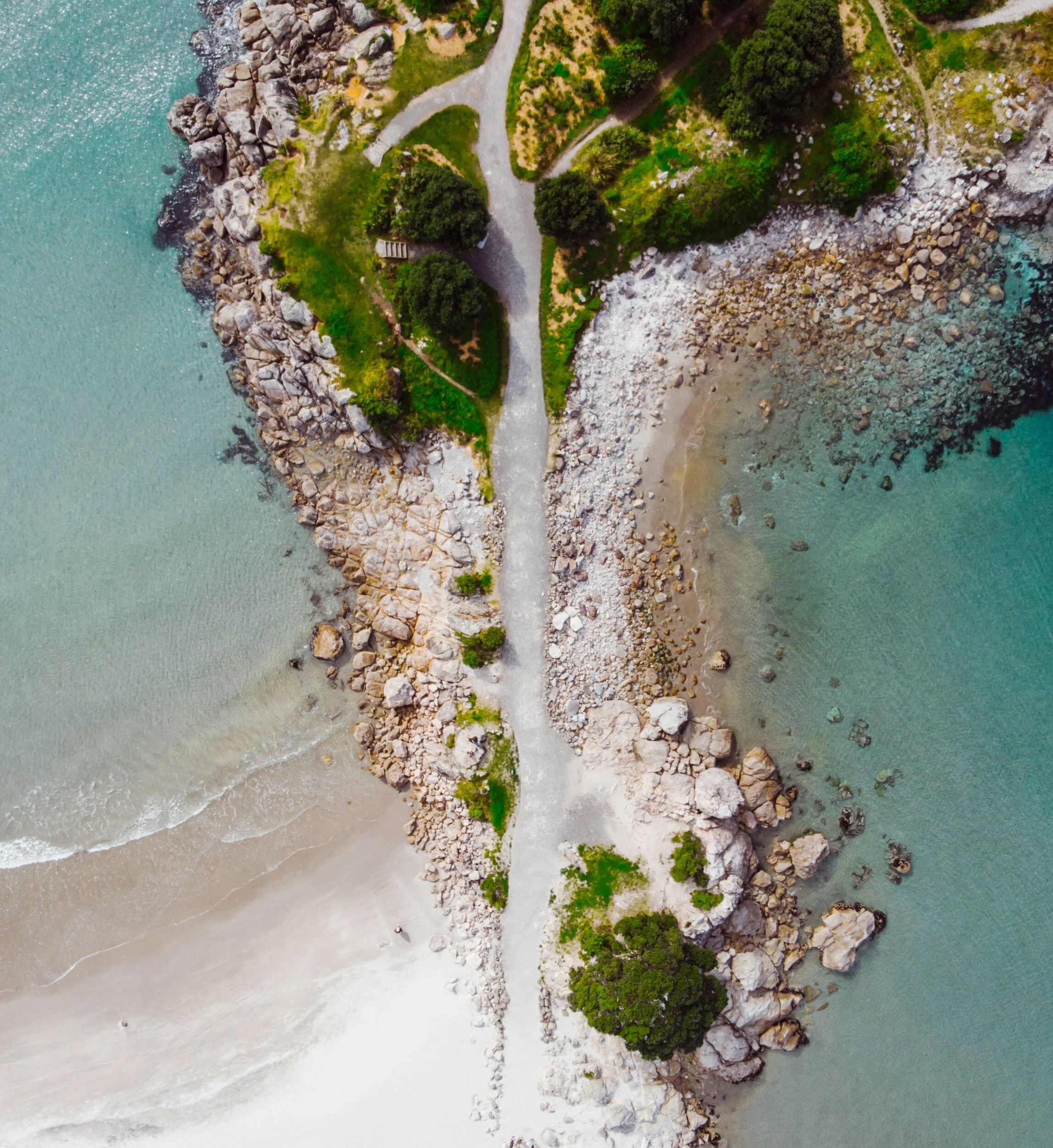 Aerial view of a coastal area with a rocky shoreline, sandy beach, and turquoise waters, with a narrow dirt path running through green grassy and wooded areas.